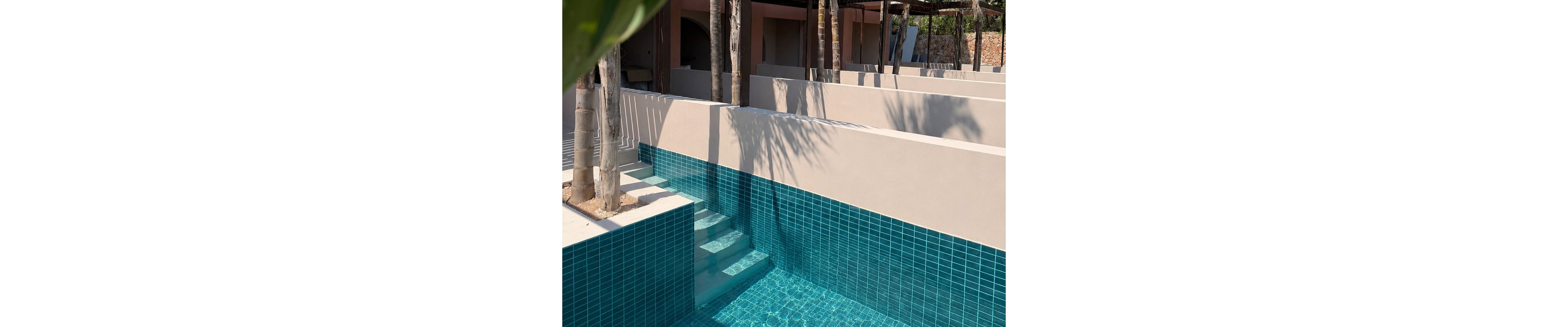 A modern outdoor pool features bright turquoise tiles and submerged steps leading into clear water. Minimalist beige walls create geometric lines and shadows around the pool area. Tall palm trees rise through openings in the deck, casting dappled shade on the surfaces. The scene suggests a tranquil, contemporary resort environment under strong sunlight.