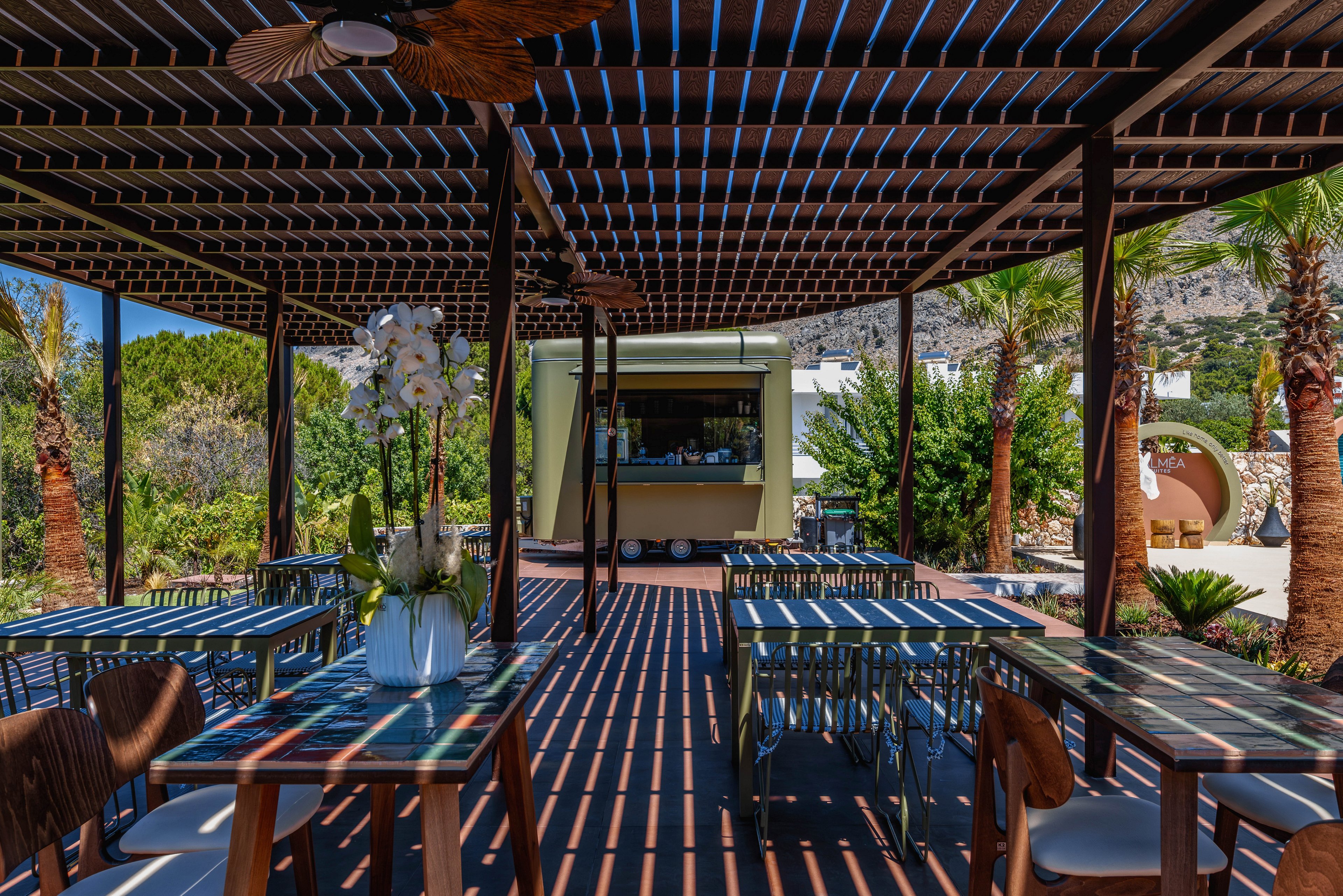 An open-air café terrace features tables and chairs arranged beneath a slatted pergola casting strong striped shadows on the floor. A small kiosk or food truck stands at the far end, framed by palm trees and lush greenery. Bright sunlight and clear blue skies create a warm, inviting atmosphere in a scenic, mountainous setting. A white floral arrangement decorates one of the foreground tables, adding a touch of elegance to the casual space.