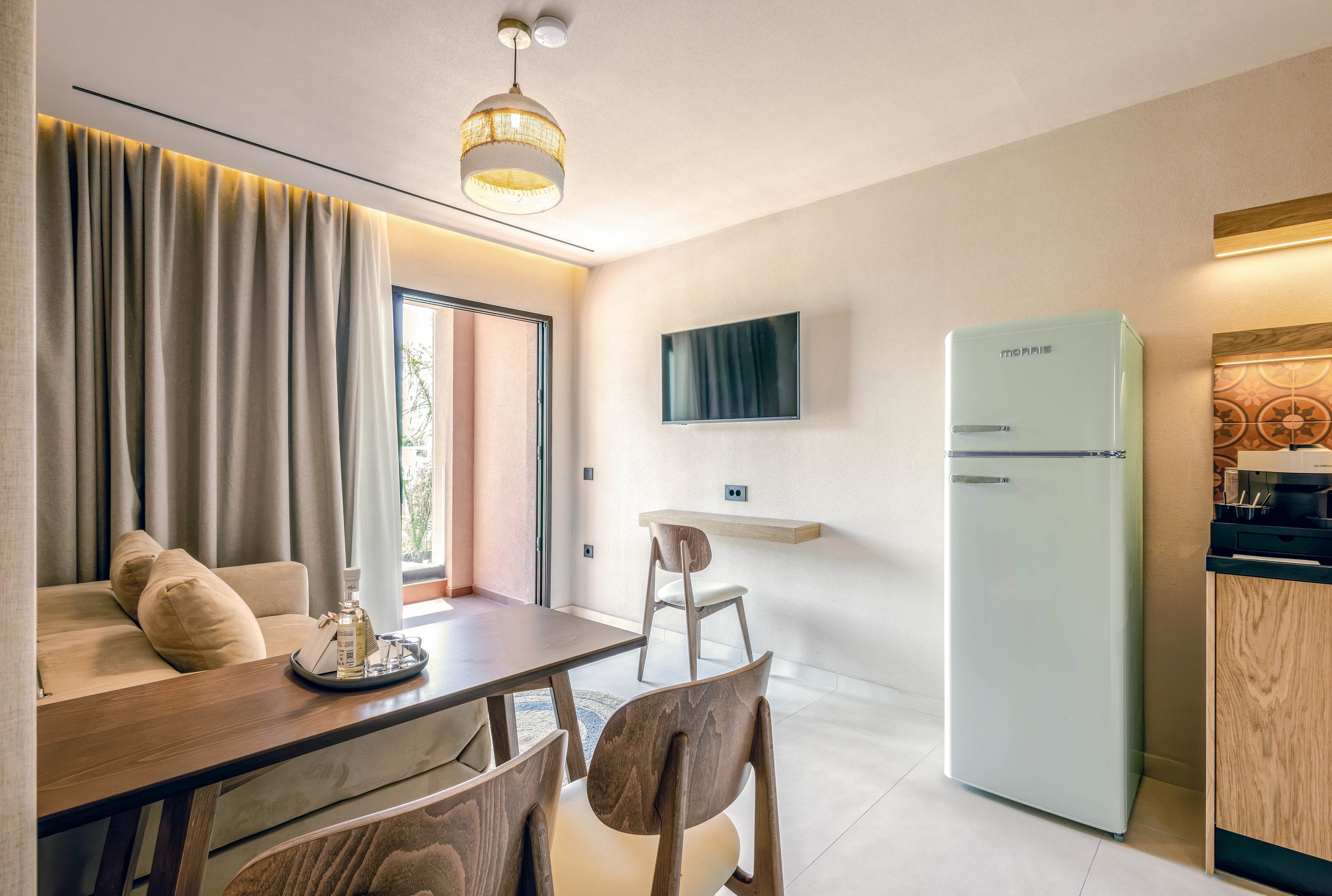 Contemporary studio interior featuring a small dining table and chairs beside a sofa. A flat-screen TV is mounted on the wall opposite a tall white refrigerator. Floor-to-ceiling curtains frame a glass door leading to a balcony with greenery outside. Warm neutral tones, soft lighting, and minimalist decor create a cozy, modern atmosphere.