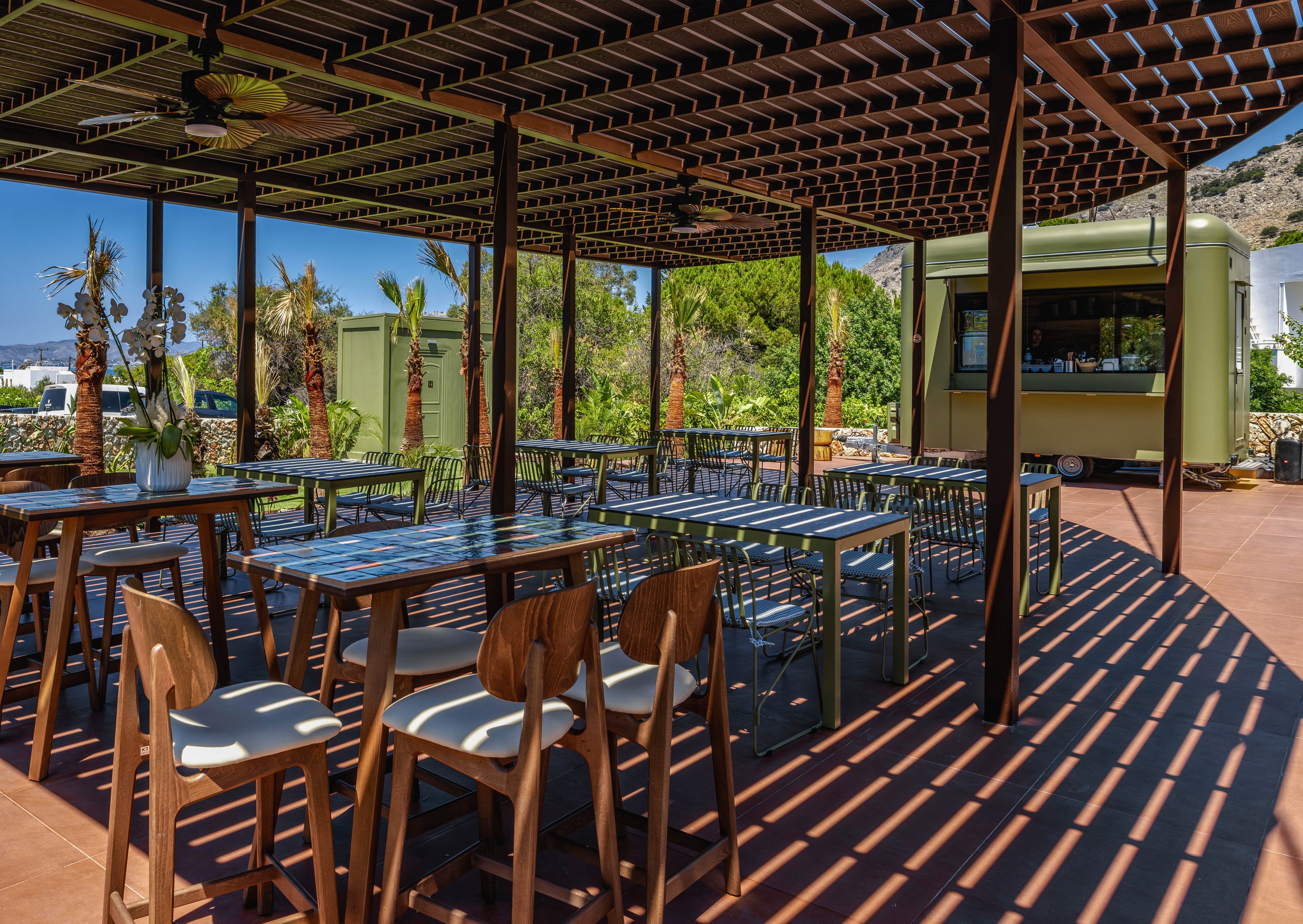 An open-air café terrace features modern wooden and metal tables and chairs under a slatted pergola. Strong sunlight creates bold striped shadows across the floor and furniture. In the background, a small kiosk or bar stands near palm trees and greenery, with hills visible beyond. The scene suggests a relaxed, warm-weather setting for dining or refreshments.