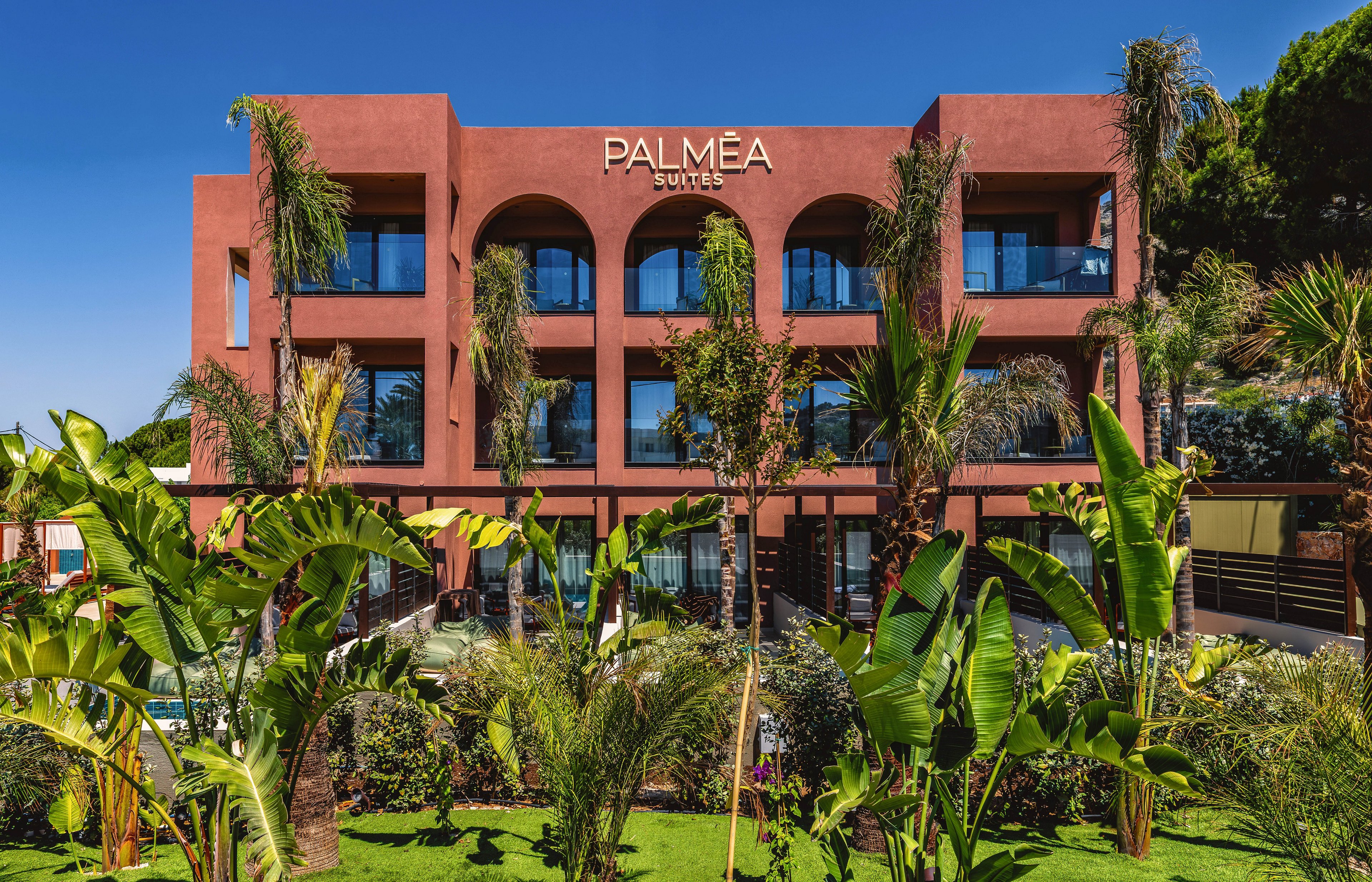 A terracotta-colored building labeled PALMEA SUITES stands behind a vibrant tropical garden. Tall palm trees and broad-leaf plants frame the view of the hotel’s arched balconies. The scene is brightly lit under a clear blue sky, suggesting a warm, sunny climate. The overall atmosphere feels inviting, resort-like, and ideal for vacation stays.