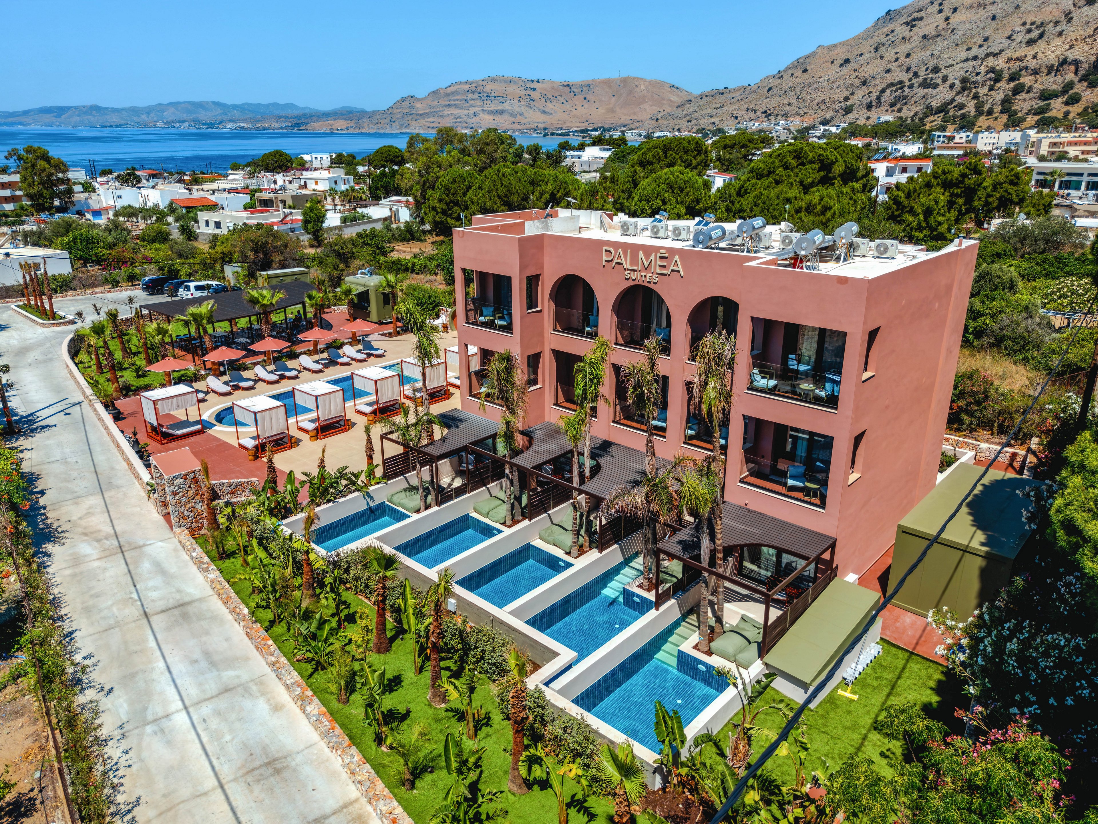 Aerial view of a modern seaside hotel named PALMIA with multiple private pools in front of the building. The property features cabana-style sunbeds and terraces surrounded by palm trees and lush landscaping. In the background, a coastal town and blue sea are visible beneath distant hills. The scene suggests a sunny, relaxing vacation atmosphere at a Mediterranean-style resort.