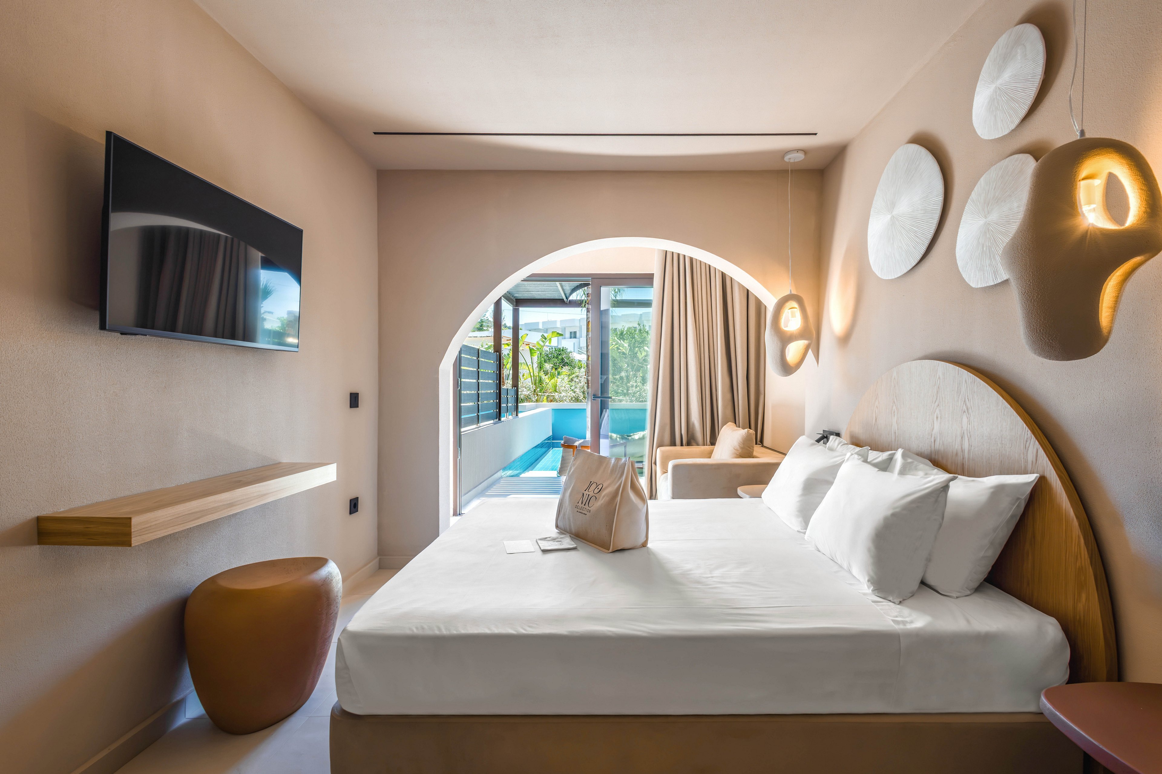 A contemporary hotel bedroom features a neatly made double bed with white linens and a beige tote bag placed on top. The room is decorated in warm neutral tones, with sculptural pendant lights and round wall art adding a stylish touch. A flat-screen TV is mounted on the wall opposite the bed, above a minimalist wooden shelf and rounded side table. Through an arched opening, sliding glass doors reveal a terrace with direct access to a private pool and lush greenery outside.