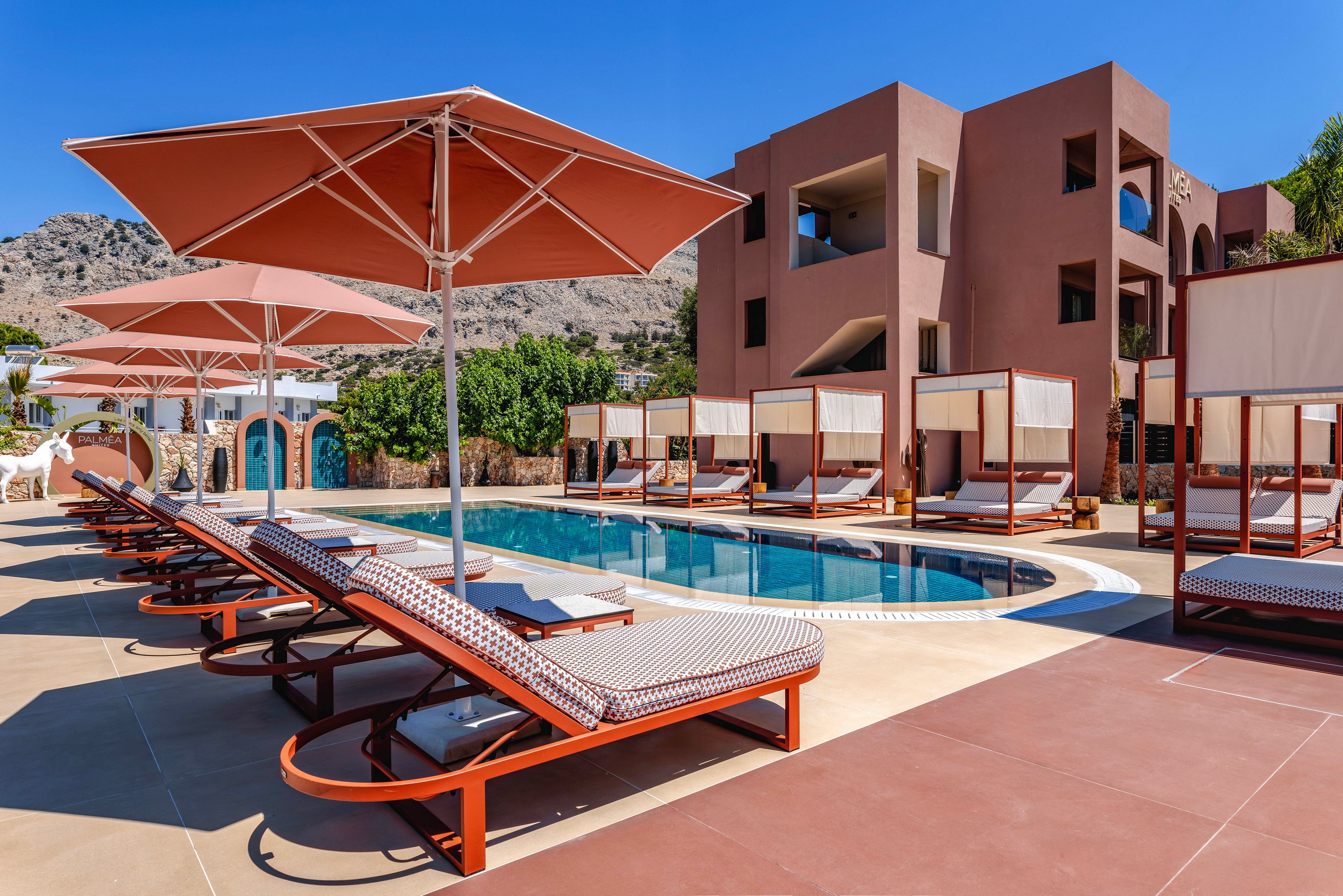 A contemporary resort pool area features rows of cushioned sun loungers under large umbrellas. Private cabanas line one side of the pool, facing a modern multi-story building with balconies. The scene is set against a backdrop of rocky mountains under a clear blue sky, suggesting a warm, sunny climate. Clean lines, neutral tones, and turquoise water create a relaxed, upscale vacation atmosphere.