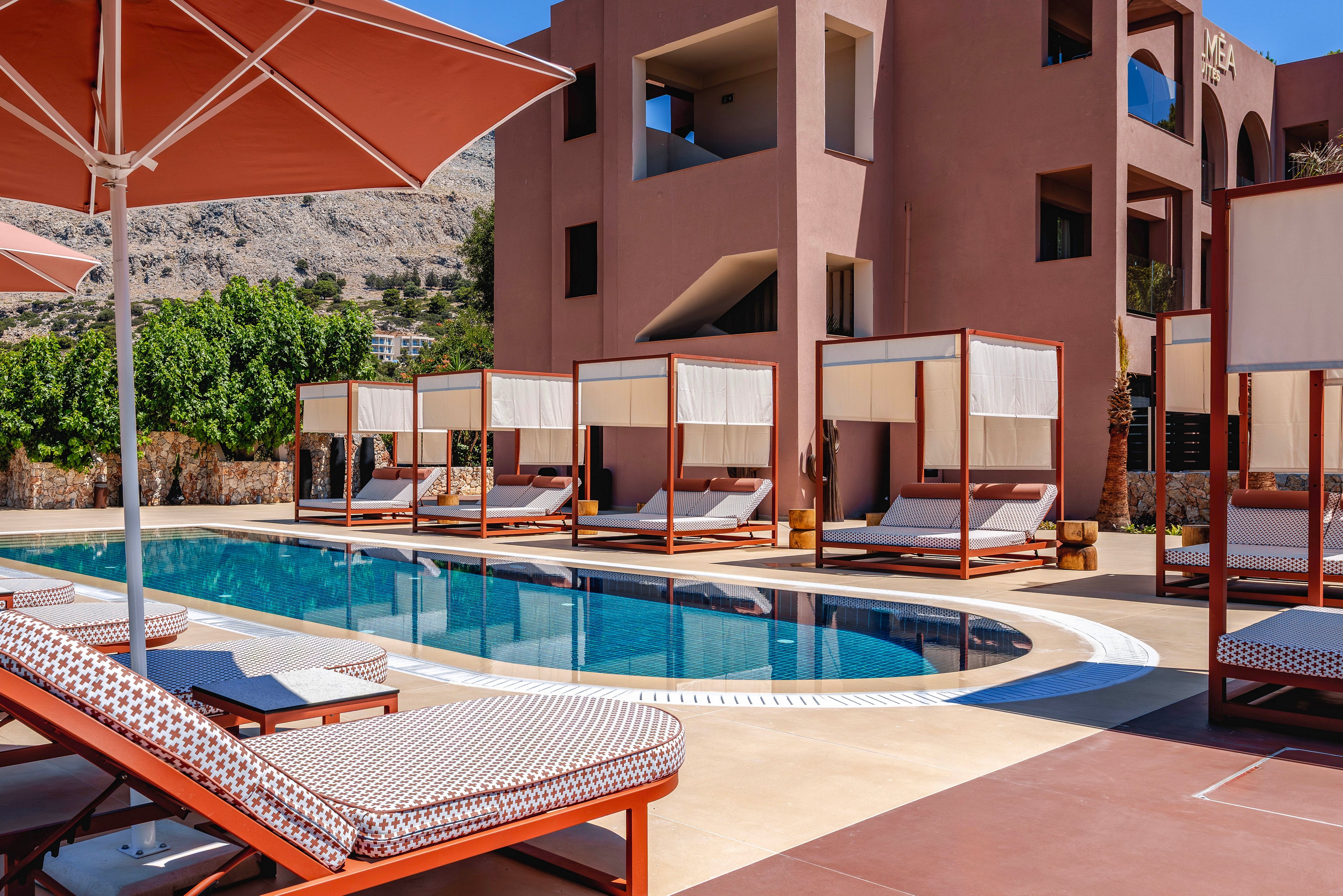 Outdoor hotel pool area with contemporary cabanas and cushioned loungers arranged along the water. A multi-story building with balconies and arched openings rises behind the pool, framed by rocky hills and greenery. Large umbrellas provide shade over patterned sunbeds, creating a relaxed vacation atmosphere. The scene is bright and colorful, suggesting a warm, sunny day at a Mediterranean-style resort.