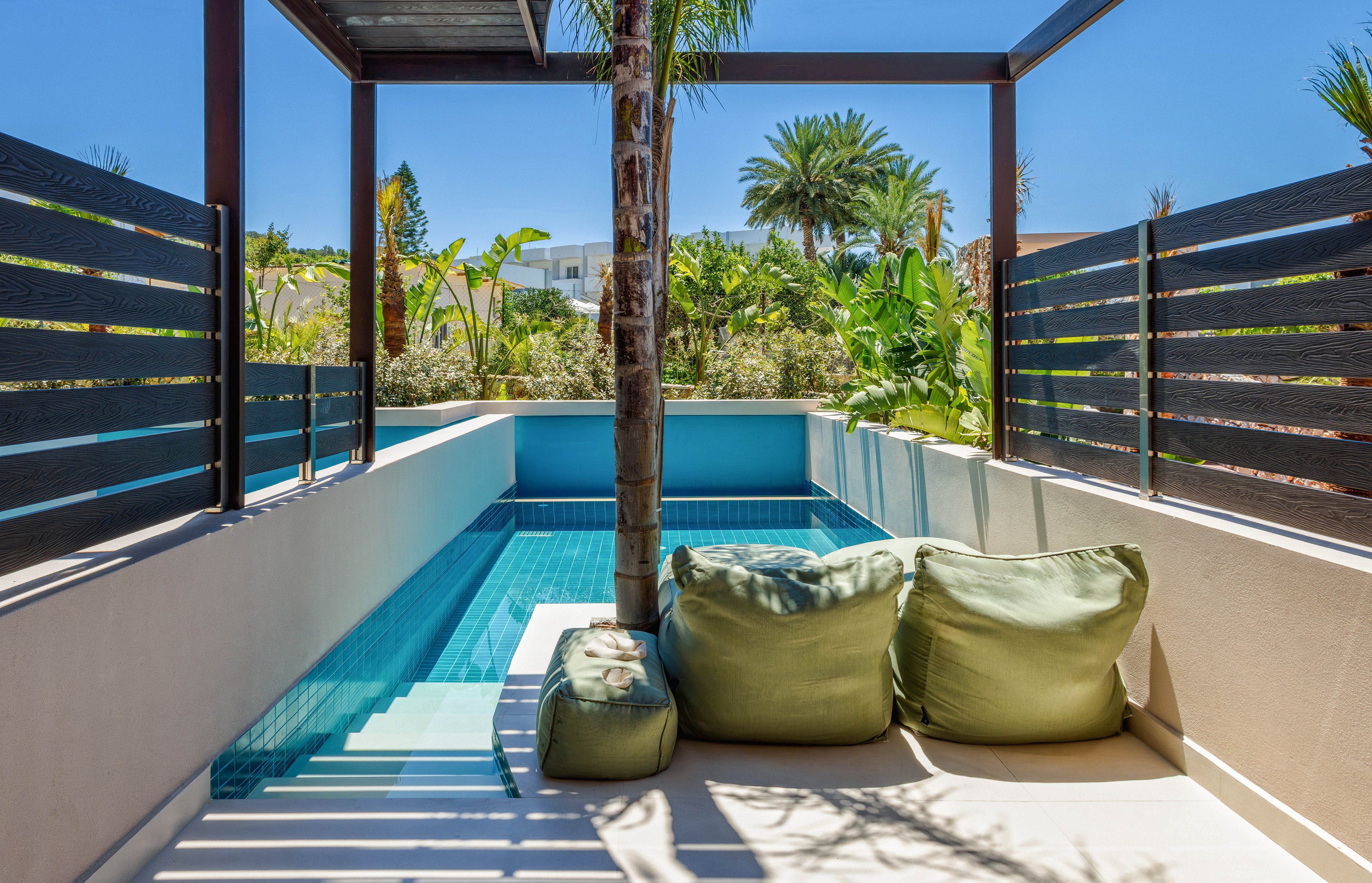 A narrow private plunge pool sits beside a small terrace with green beanbag loungers. Dark horizontal slat fencing frames the space, while tropical plants and palm trees surround the area. Bright blue water reflects the clear sunny sky, creating a relaxing vacation atmosphere. Contemporary architecture and clean lines emphasize a modern outdoor retreat.