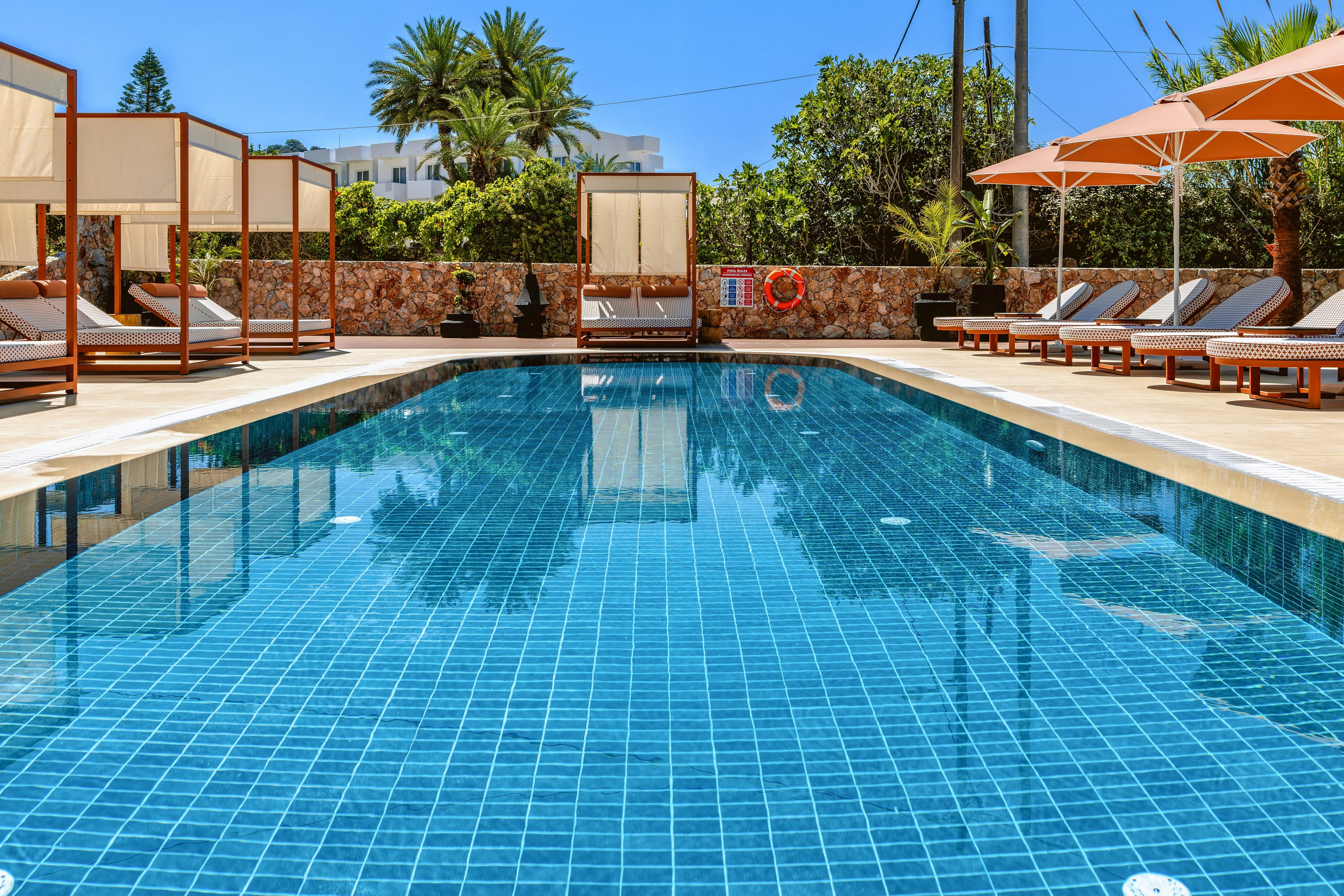 A clear blue tiled swimming pool stretches between rows of sun loungers and cabanas at a resort. White cushions and umbrellas line the deck, creating a comfortable space for relaxation. Palm trees and lush greenery rise behind a stone wall, with white buildings visible in the background. Bright daylight and vivid colors give the scene a warm, inviting vacation atmosphere.