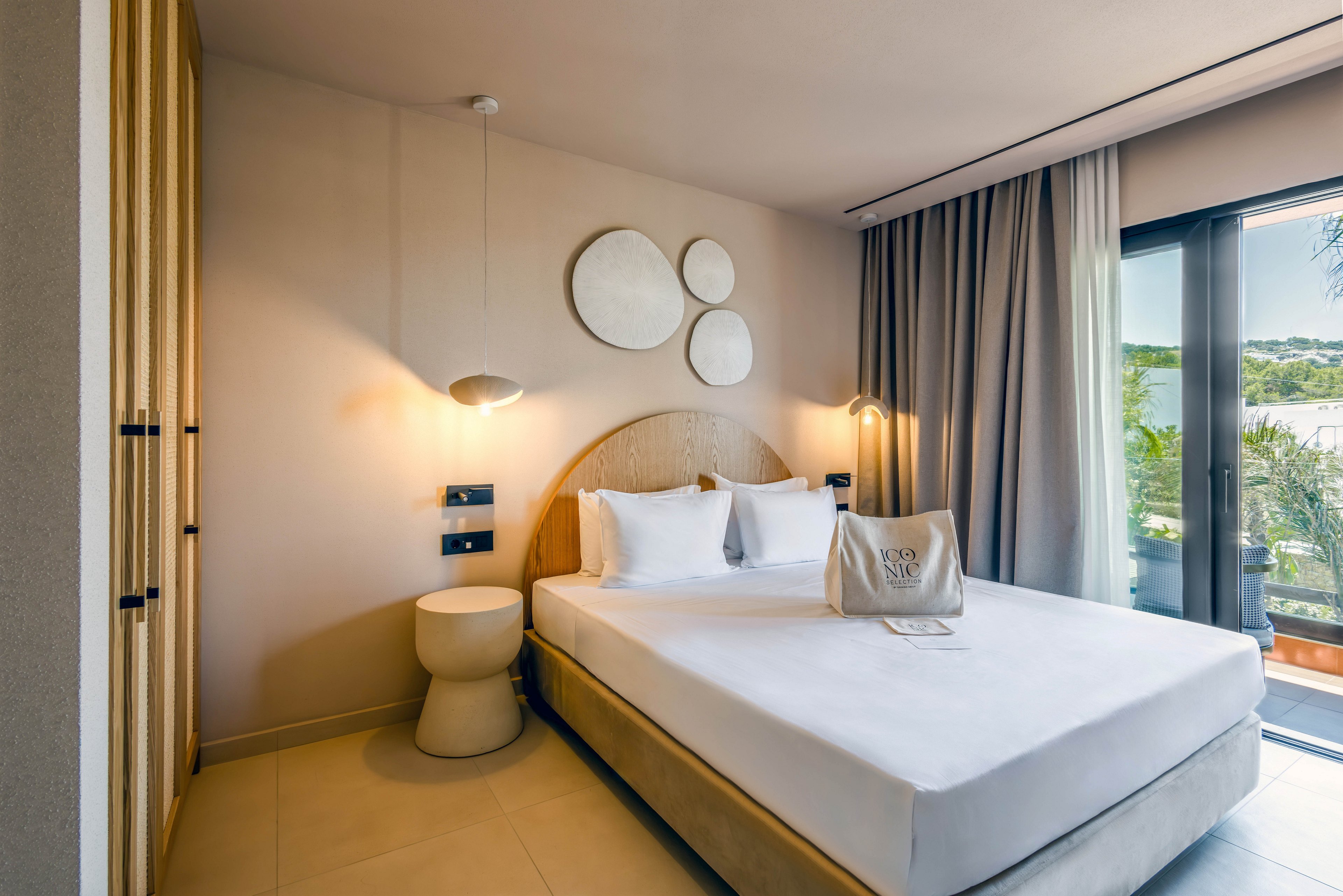 A contemporary hotel bedroom features a neatly made double bed with white linens and pillows. Minimalist decor includes round wall art, a small bedside table, and hanging lamps that create a warm, inviting atmosphere. Floor-to-ceiling curtains frame a glass door leading to a balcony with a scenic outdoor view. A neutral color palette of beige and soft tones enhances the calm, relaxing mood of the space.