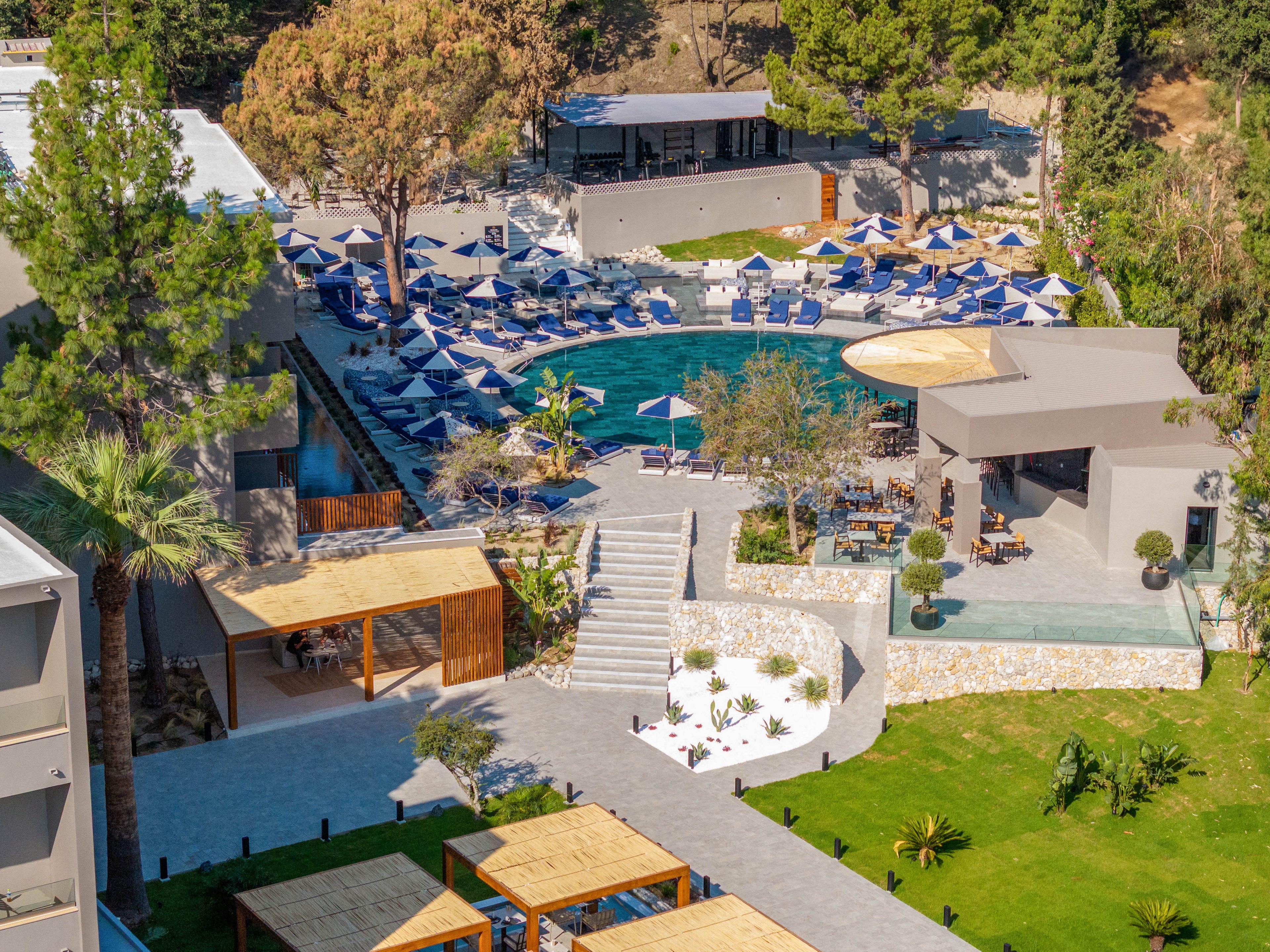 A resort-style outdoor pool area is surrounded by numerous blue sun loungers and umbrellas. Modern buildings and terraces frame the pool, with stairs leading down to landscaped paths and seating areas. Green lawns, trees, and shrubs add a lush feel to the sunny setting. The scene suggests a relaxing vacation atmosphere at a contemporary hotel or resort.
