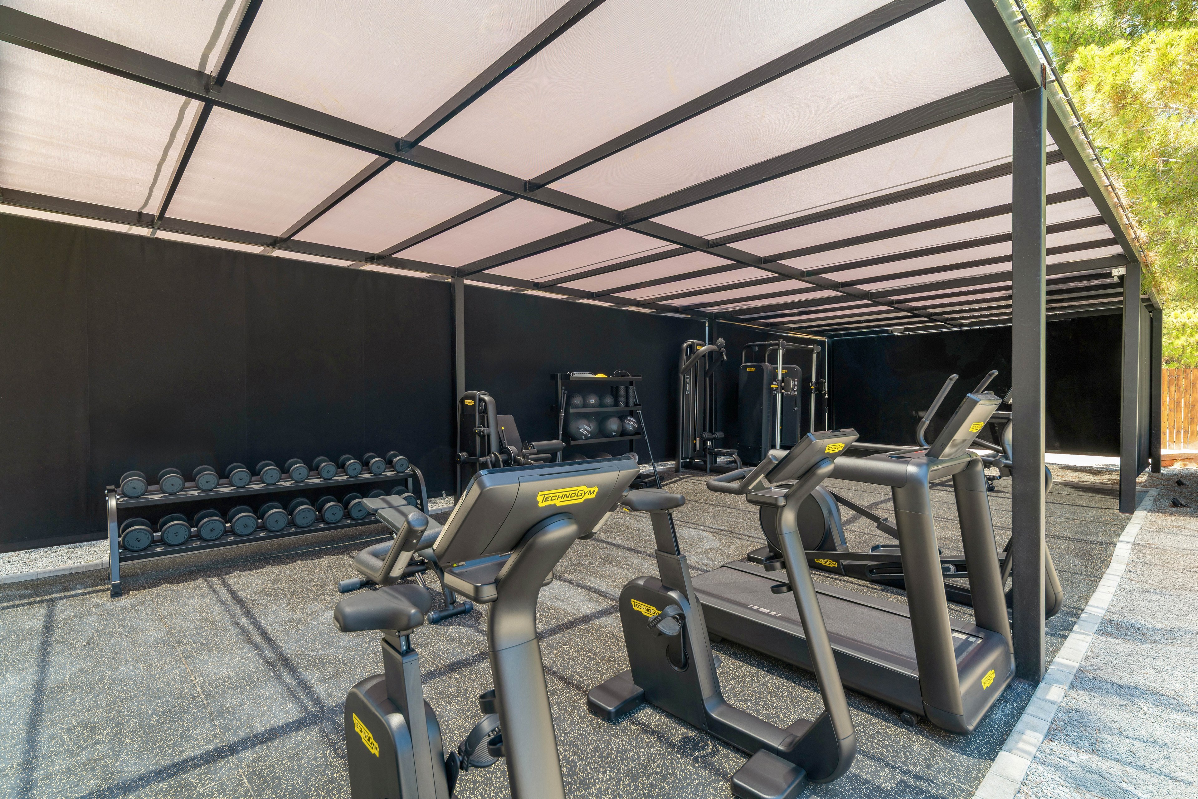 An open-air gym features several treadmills and strength machines arranged under a covered structure. A rack of dumbbells lines one side of the space, providing free weights for strength training. The area has a clean, minimalist design with neutral tones and ample natural light. No people are present, emphasizing the equipment and spacious layout.
