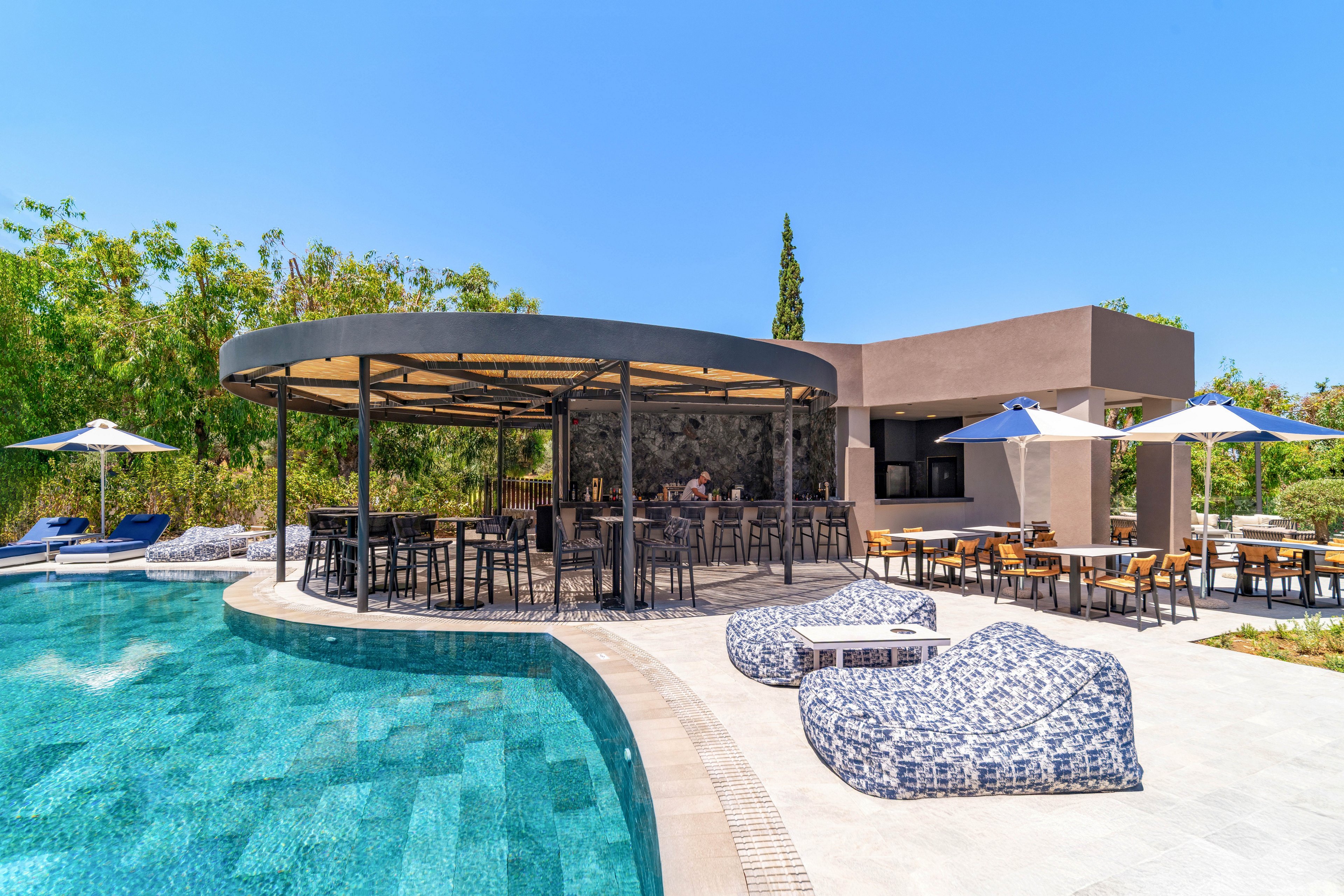A contemporary outdoor bar sits beside a clear blue swimming pool in a sunny resort setting. High stools surround the circular bar structure while tables and chairs extend into a shaded dining area. Patterned lounge chairs and umbrellas line the poolside, inviting guests to relax. Lush greenery and a bright blue sky frame the tranquil leisure scene.
