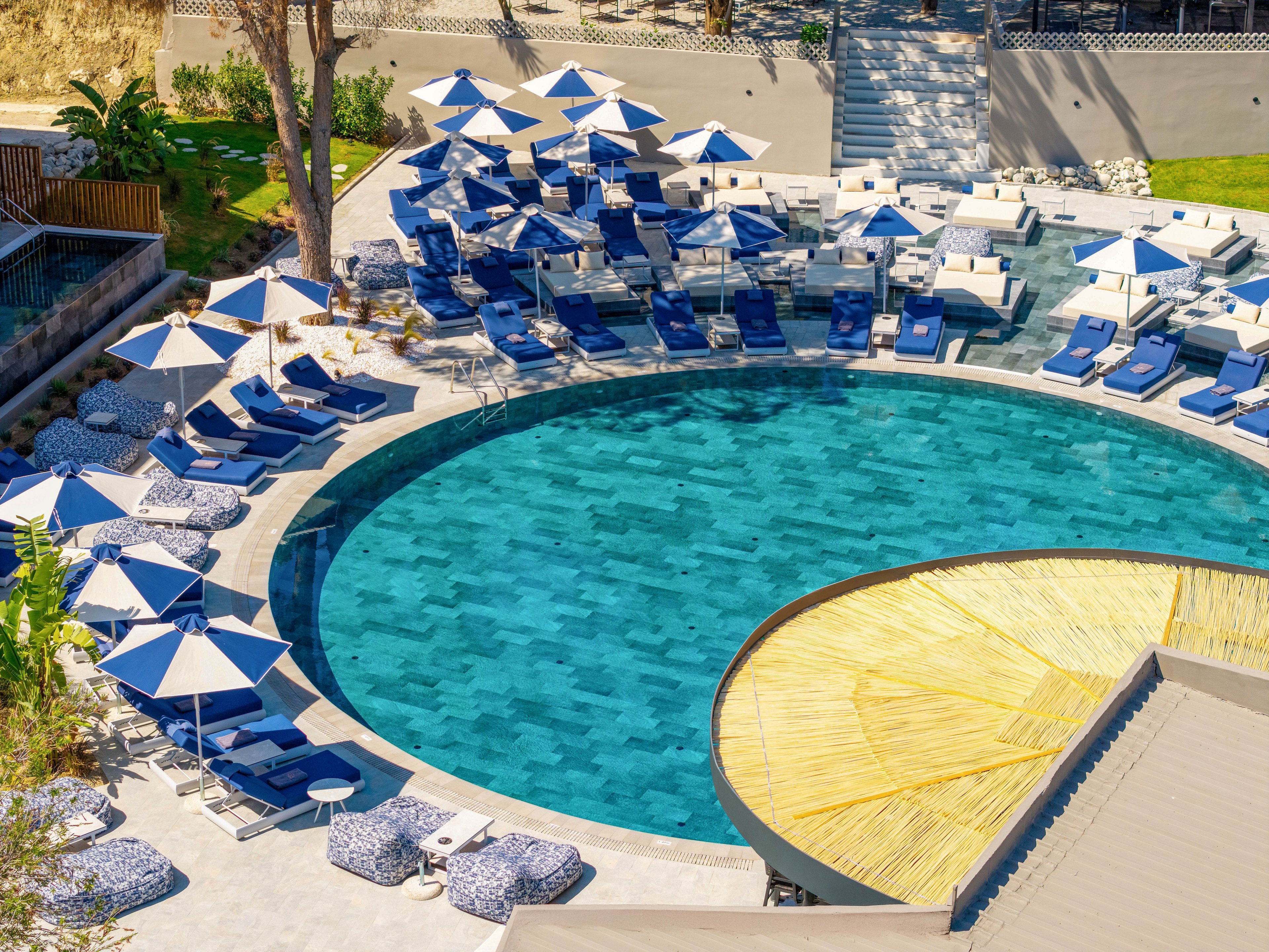 A circular resort swimming pool is surrounded by numerous blue lounge chairs and white-and-blue umbrellas. The scene is viewed from above, highlighting the clear turquoise water and the geometric layout of the pool deck. A thatched-roof structure partially covers one side of the pool, adding a tropical touch. The bright sunlight and clean design create a relaxing vacation atmosphere.