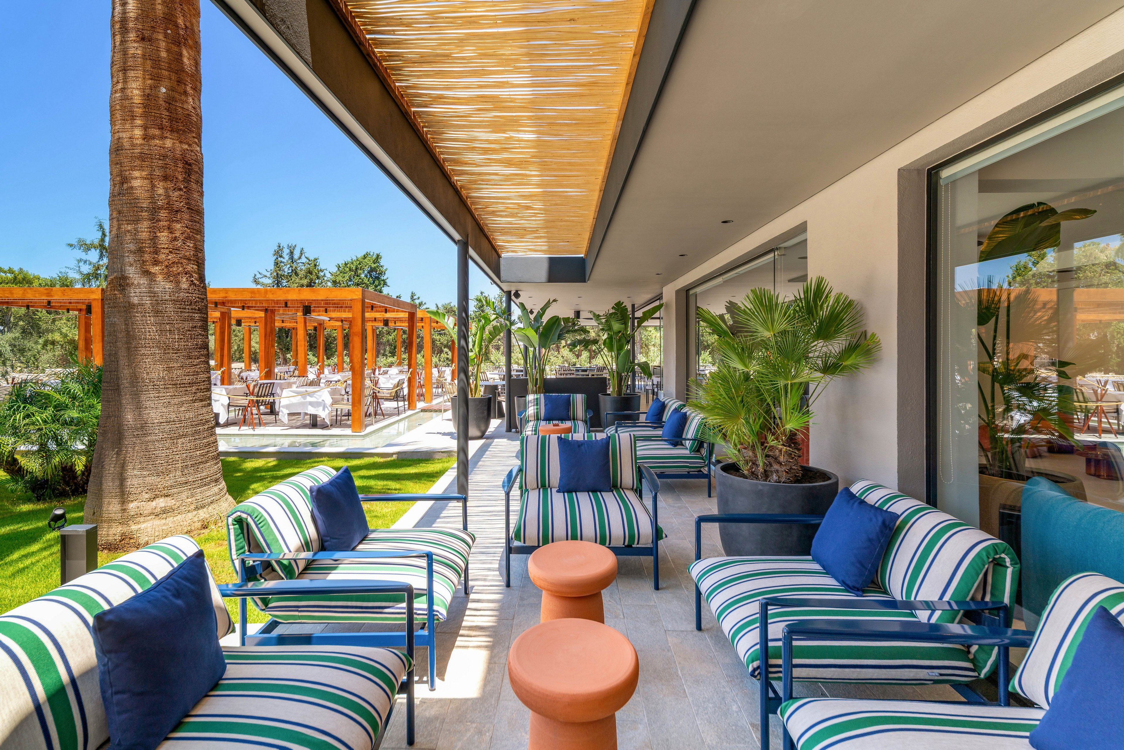 A contemporary outdoor lounge area features striped cushioned chairs and small round tables arranged along a covered patio. The space overlooks a lush garden with palm trees and manicured grass, creating a resort-like atmosphere. A wooden pergola structure with dining tables is visible in the background, suggesting an adjacent restaurant or event space. Large windows and potted plants enhance the airy, relaxed feel of this modern hospitality setting.