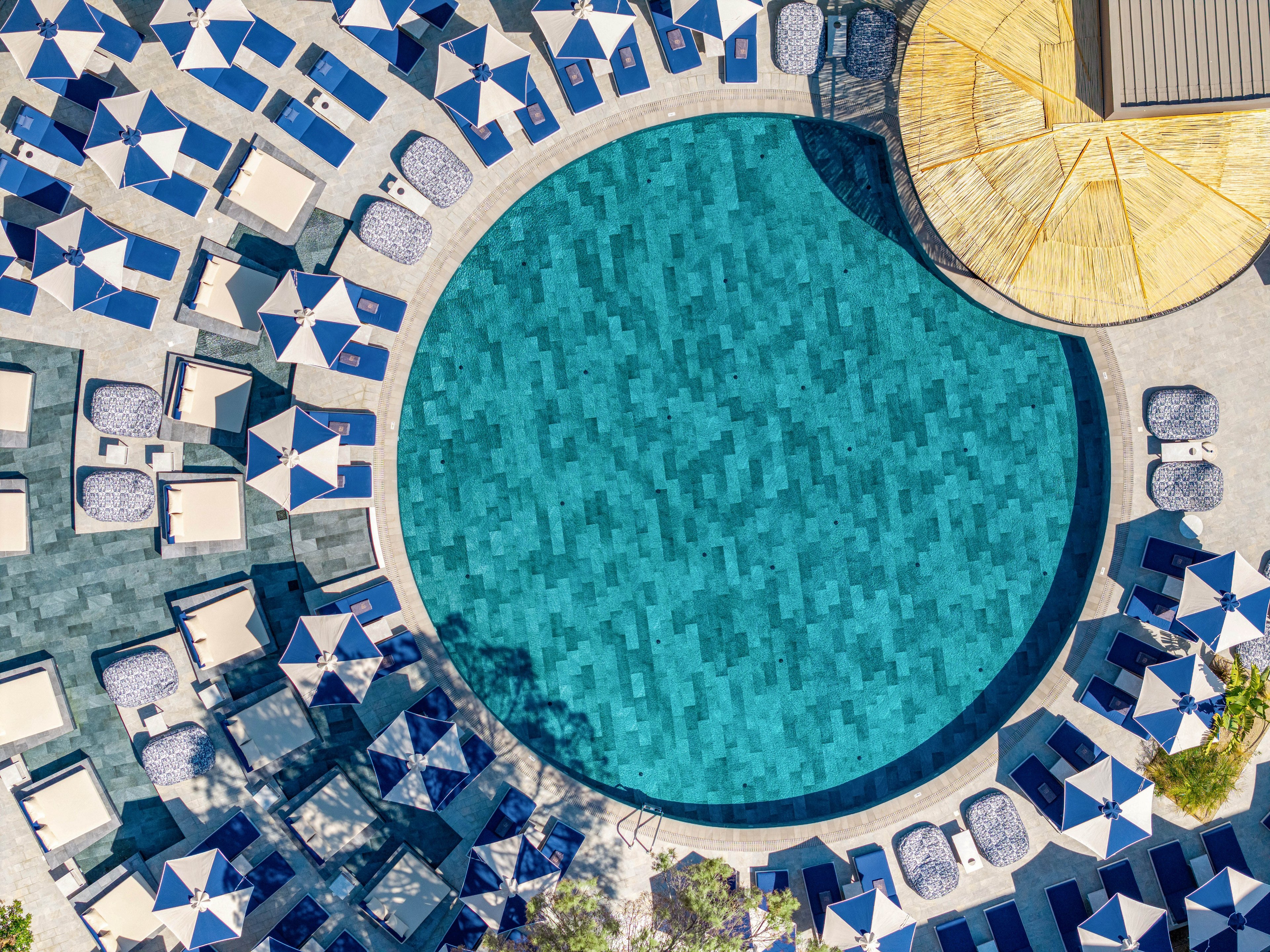 An aerial view shows a large circular swimming pool surrounded by neatly arranged lounge chairs and blue-and-white umbrellas. The pool’s tiled surface creates a mosaic of varying blue tones, giving the water a rich, textured appearance. Sun loungers and umbrellas form a geometric pattern around the pool, suggesting a well-organized resort or hotel setting. A thatched or wooden-roofed structure partially frames the scene, adding a warm contrast to the cool blue palette.