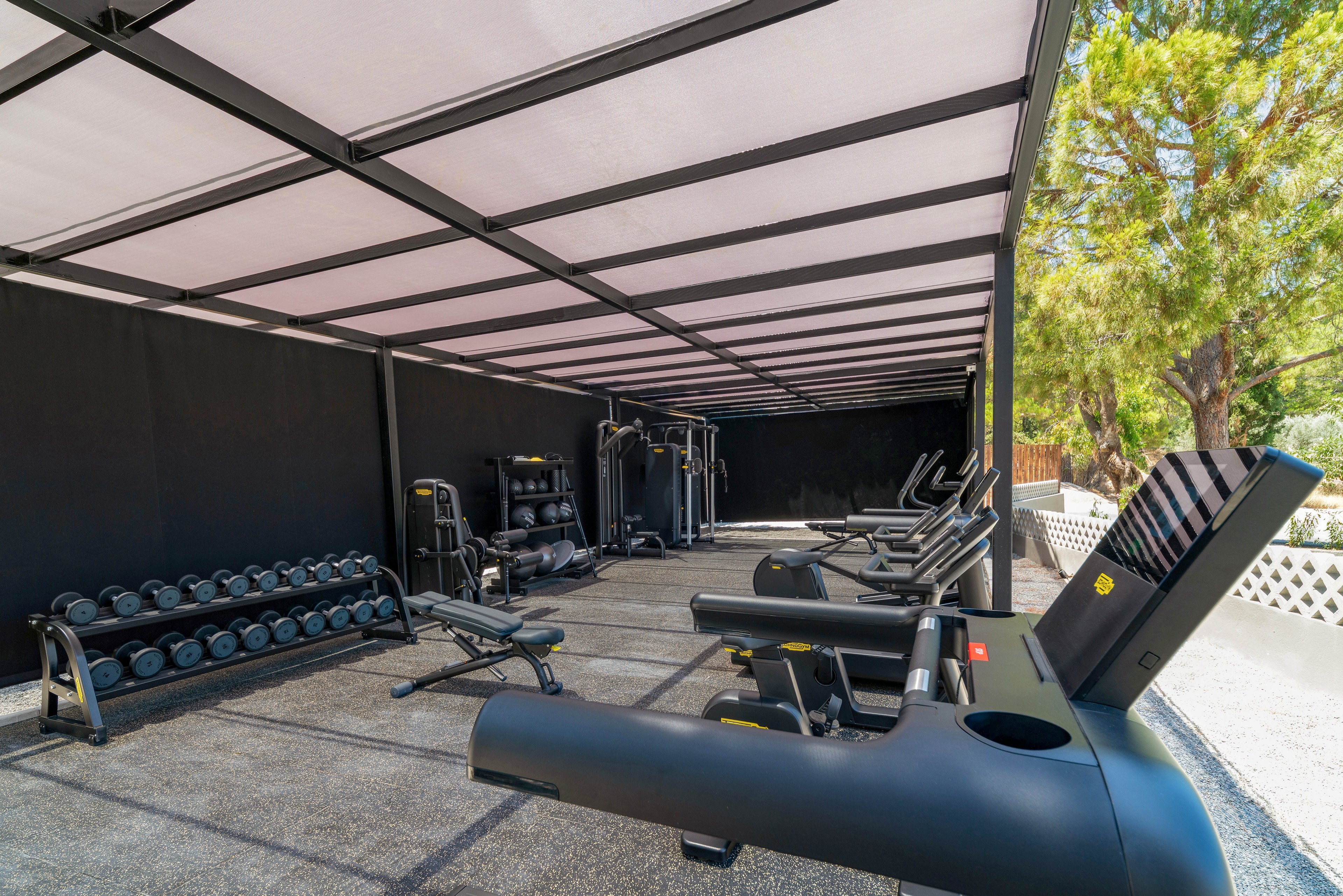 An open-air gym features a variety of modern fitness machines under a covered structure. Several treadmills and stationary bikes line the right side, while weight benches and dumbbell racks occupy the left. The space has a clean, minimalist design with black equipment and a light roof panel. Trees and natural greenery are visible beyond the gym, creating a bright and airy workout environment.