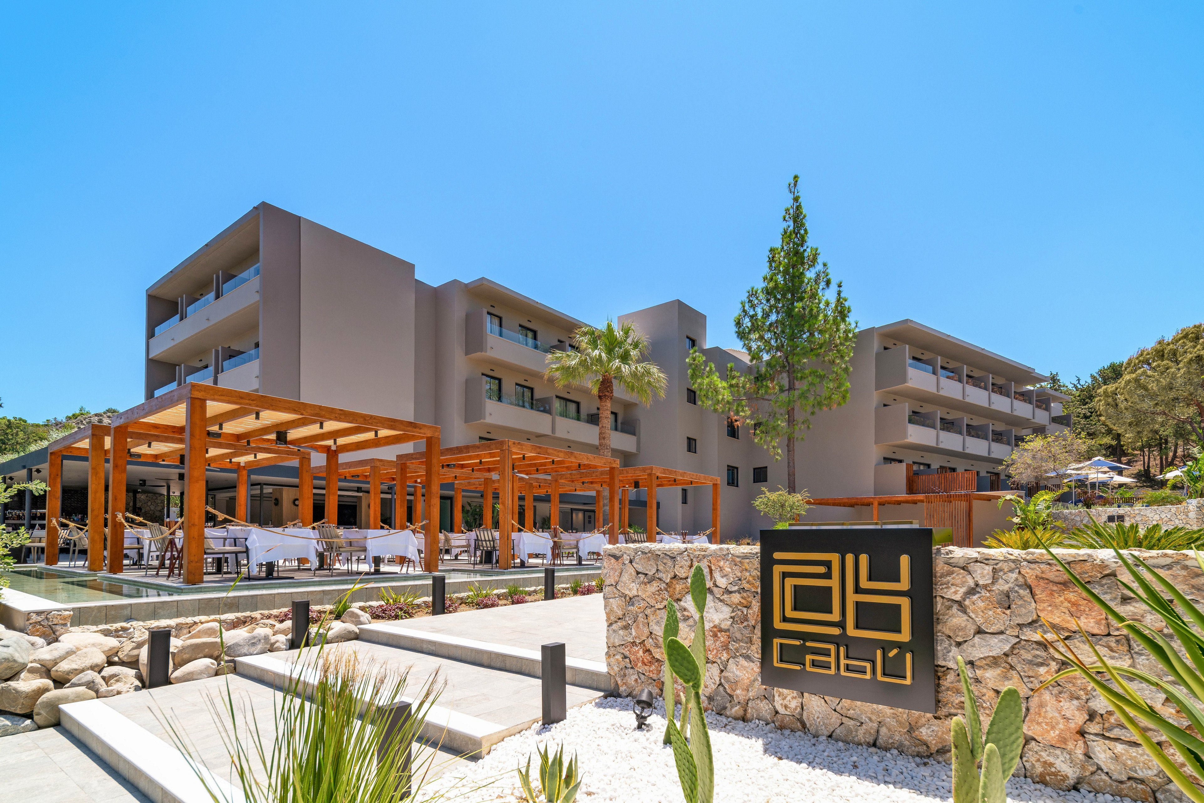 A contemporary multi-story resort hotel stands under a clear blue sky. In the foreground, a stylish outdoor dining terrace with wooden pergolas extends along the front of the building. A landscaped entrance with stone walls, white gravel, and green plants leads toward the hotel. A dark sign with geometric symbols and the word 'cabu' is prominently displayed near the pathway. The overall scene suggests a sunny, relaxed vacation atmosphere in a warm climate.