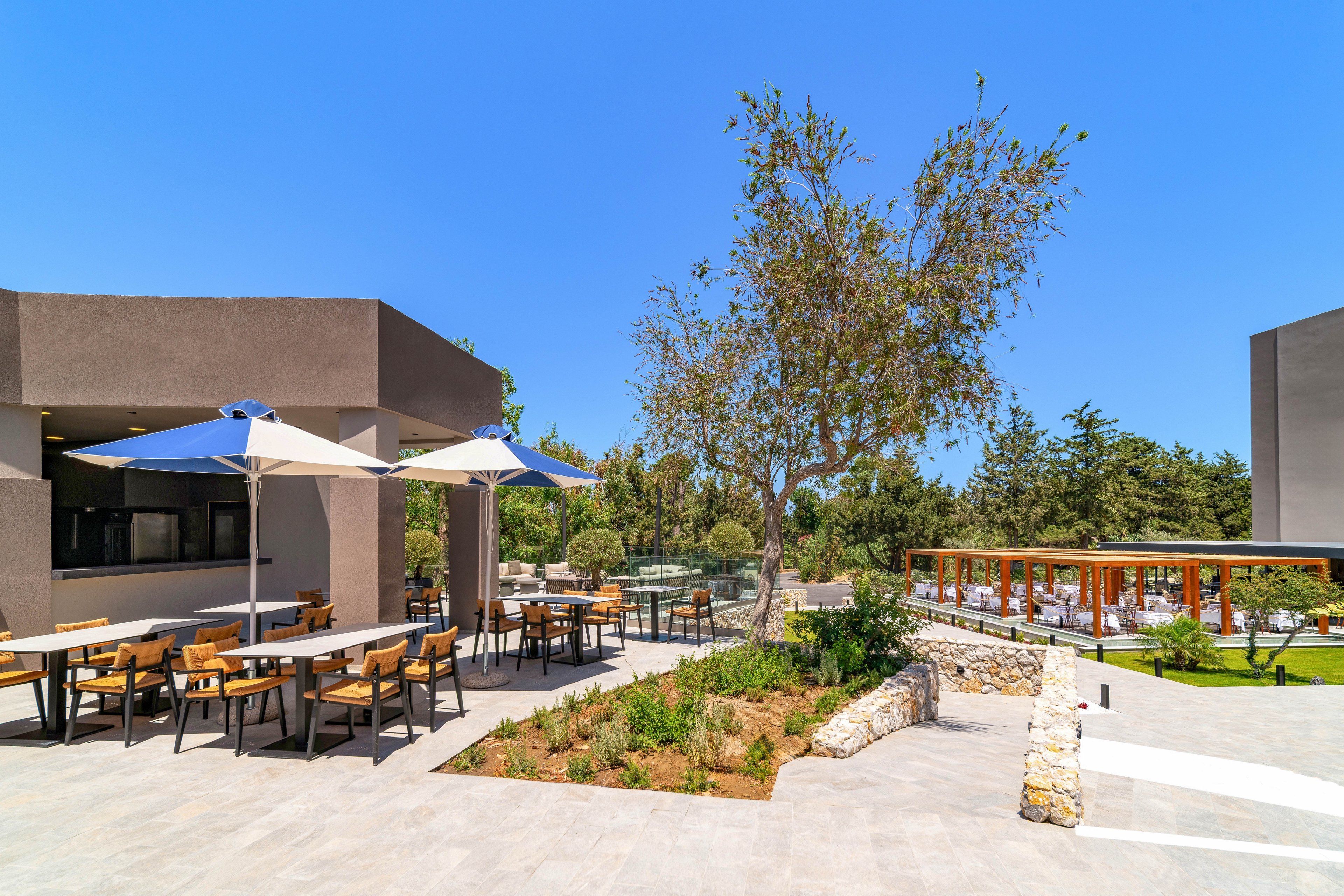 A contemporary outdoor cafe terrace features wooden tables and chairs arranged under large white and blue umbrellas. The setting is a sunny day with clear blue skies, surrounded by landscaped greenery and trees. A modern building with clean lines borders the patio, while a glass-walled structure and lawn area appear in the background. The scene conveys a relaxed, resort-like atmosphere ideal for outdoor dining and socializing.