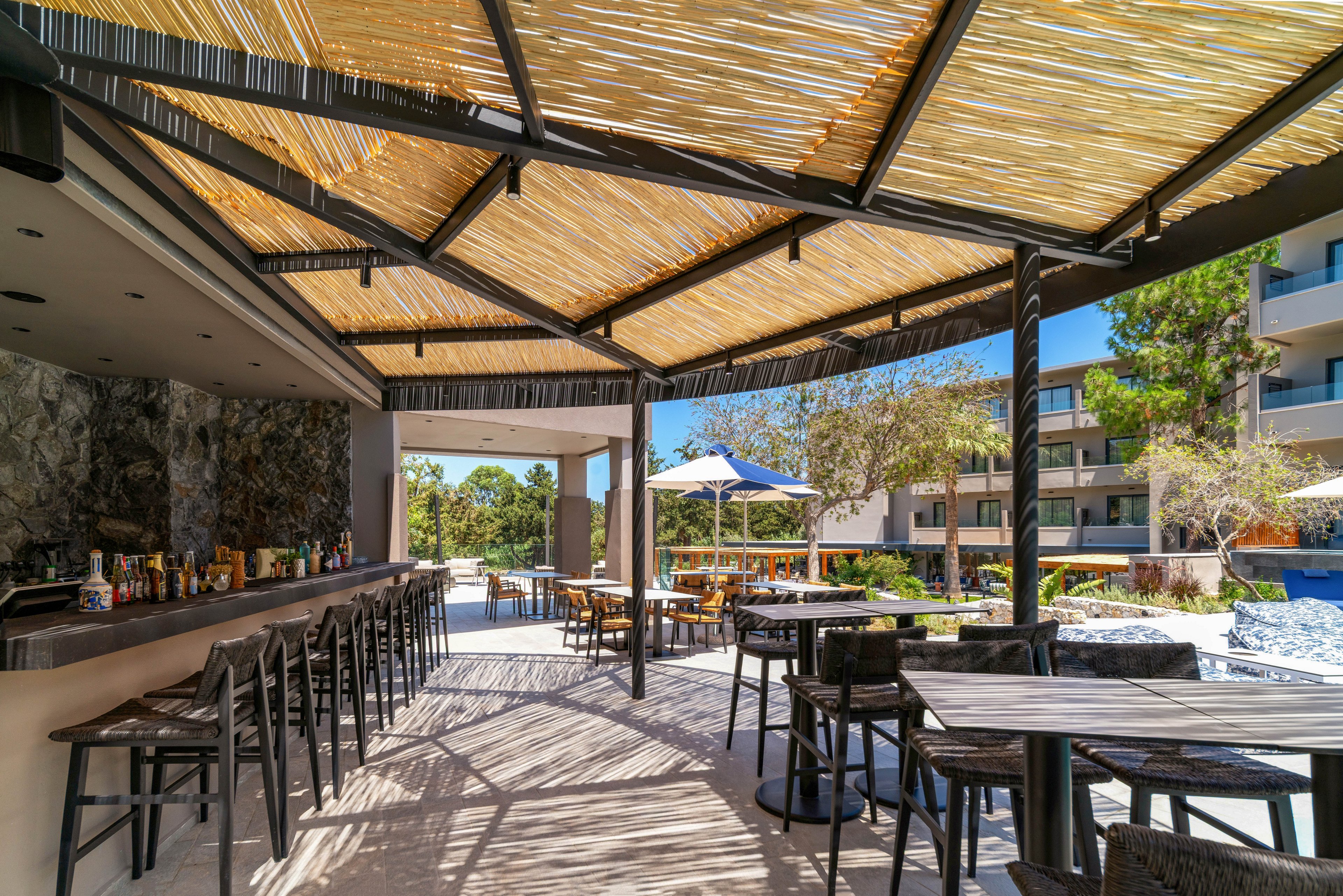 An open-air bar terrace features a long counter with barstools and various bottles arranged along the back wall. The space is shaded by a slatted wooden pergola that filters sunlight onto the tiled floor. Several tables and chairs are set up for guests, overlooking a landscaped pool area with umbrellas and modern resort buildings. The scene suggests a relaxed, vacation atmosphere in a warm, sunny climate.