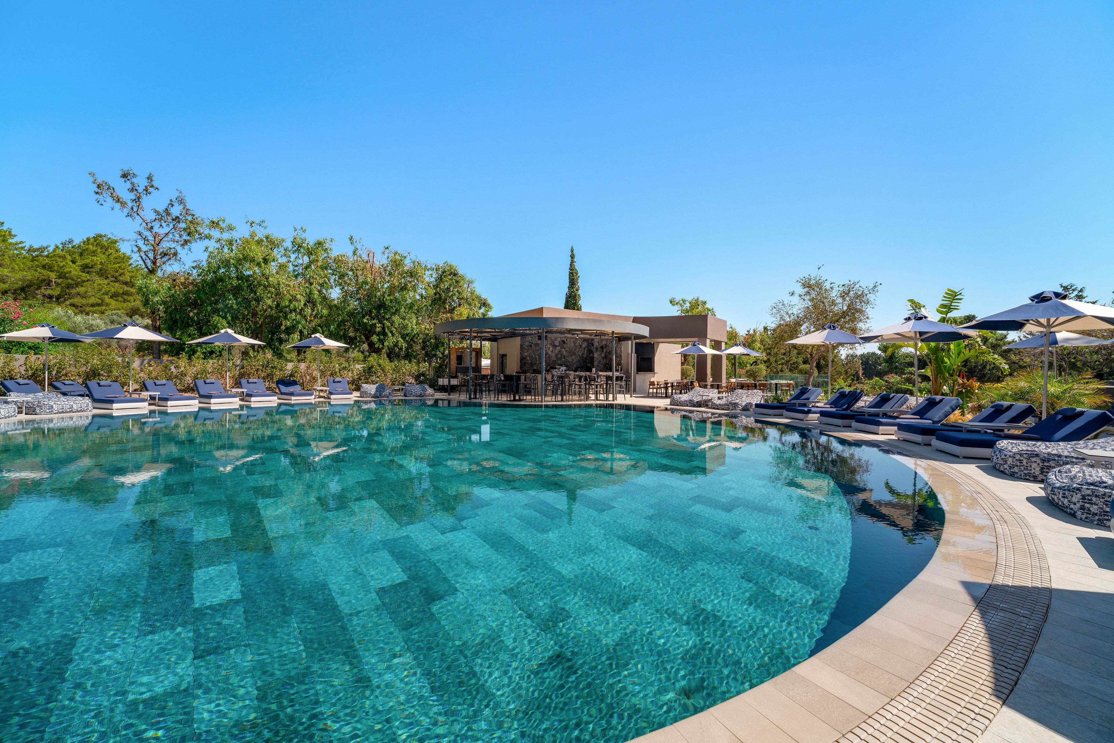 A large, clear blue resort swimming pool dominates the foreground, reflecting the bright sky above. Numerous cushioned sun loungers and umbrellas line the curved poolside, suggesting a relaxing vacation setting. A poolside bar or lounge structure sits at the far end, surrounded by lush greenery and trees. The scene is calm and inviting, with modern outdoor furniture and a clean, contemporary design.