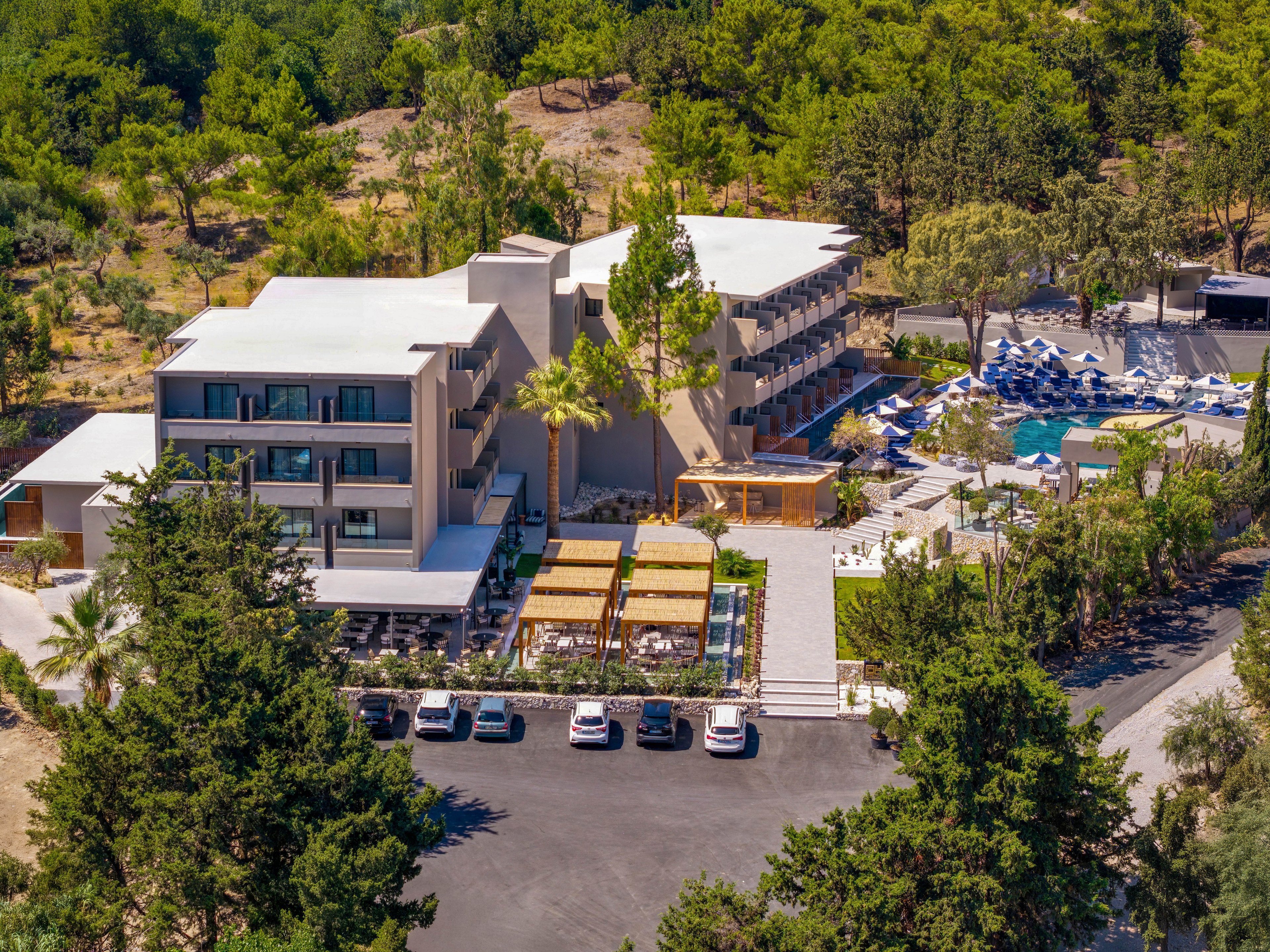 An aerial view shows a modern multi-story resort hotel nestled on a green hillside. The property features several terraced buildings with balconies facing a central courtyard. A swimming pool area with numerous blue and white sun loungers is visible on one side. Cars are parked in a paved lot at the front entrance, while dense trees and natural landscape surround the complex. The overall scene suggests a tranquil vacation destination in a warm climate.
