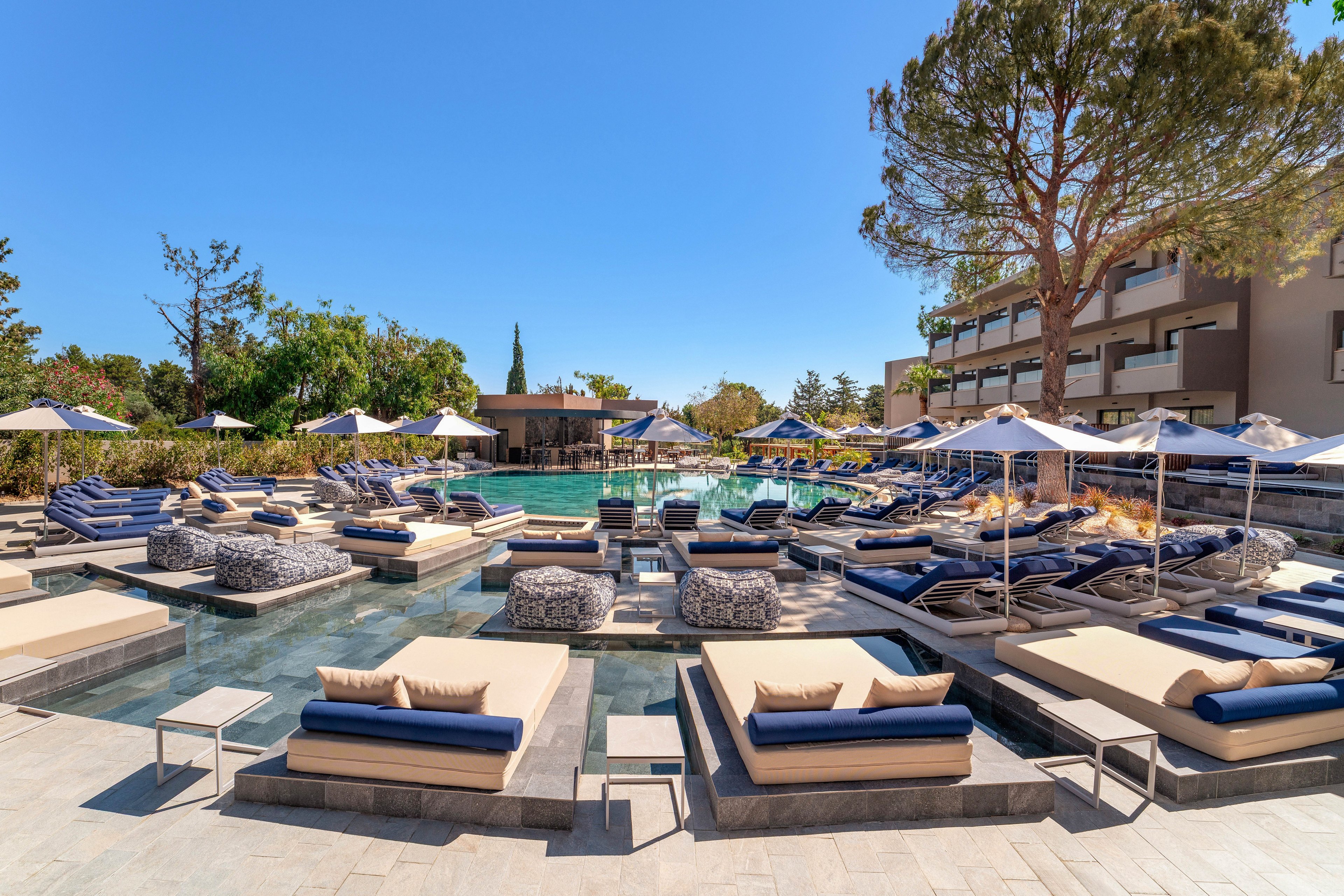 A modern resort pool area features numerous cushioned sun loungers arranged on platforms over shallow water. Blue and beige daybeds and umbrellas create a coordinated, upscale look around the pool. Trees and landscaped greenery surround the space, with a multi-story hotel building in the background. The bright daylight and clear sky emphasize a relaxed, vacation atmosphere.