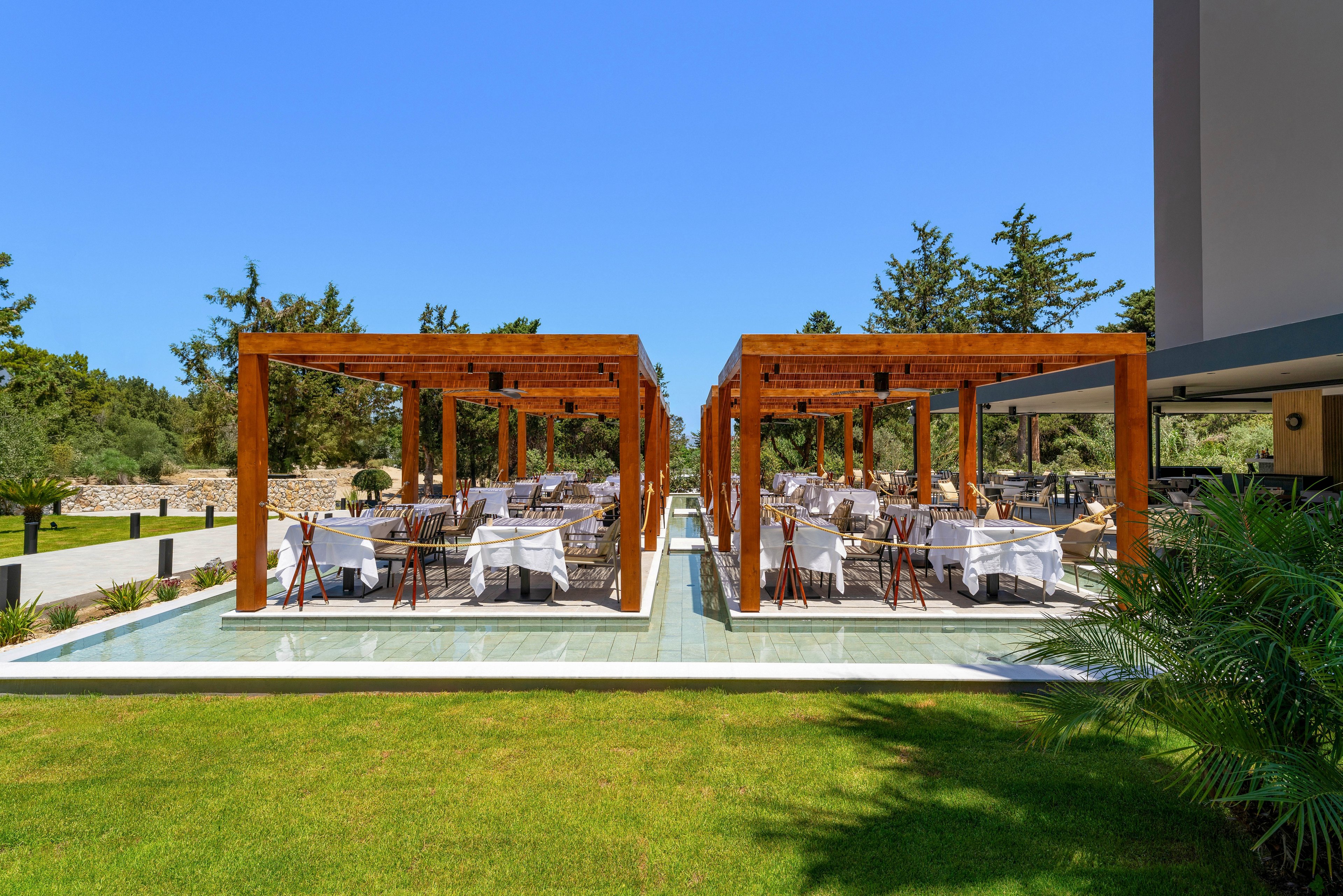 An elegant outdoor dining area is arranged beneath two parallel wooden pergolas in a landscaped resort courtyard. White tablecloths and neatly set tables create a formal restaurant atmosphere under the open sky. The pergolas stand over a reflective surface bordered by bright green grass and tropical plants. Tall trees and a clear blue sky frame the tranquil, sunny setting.