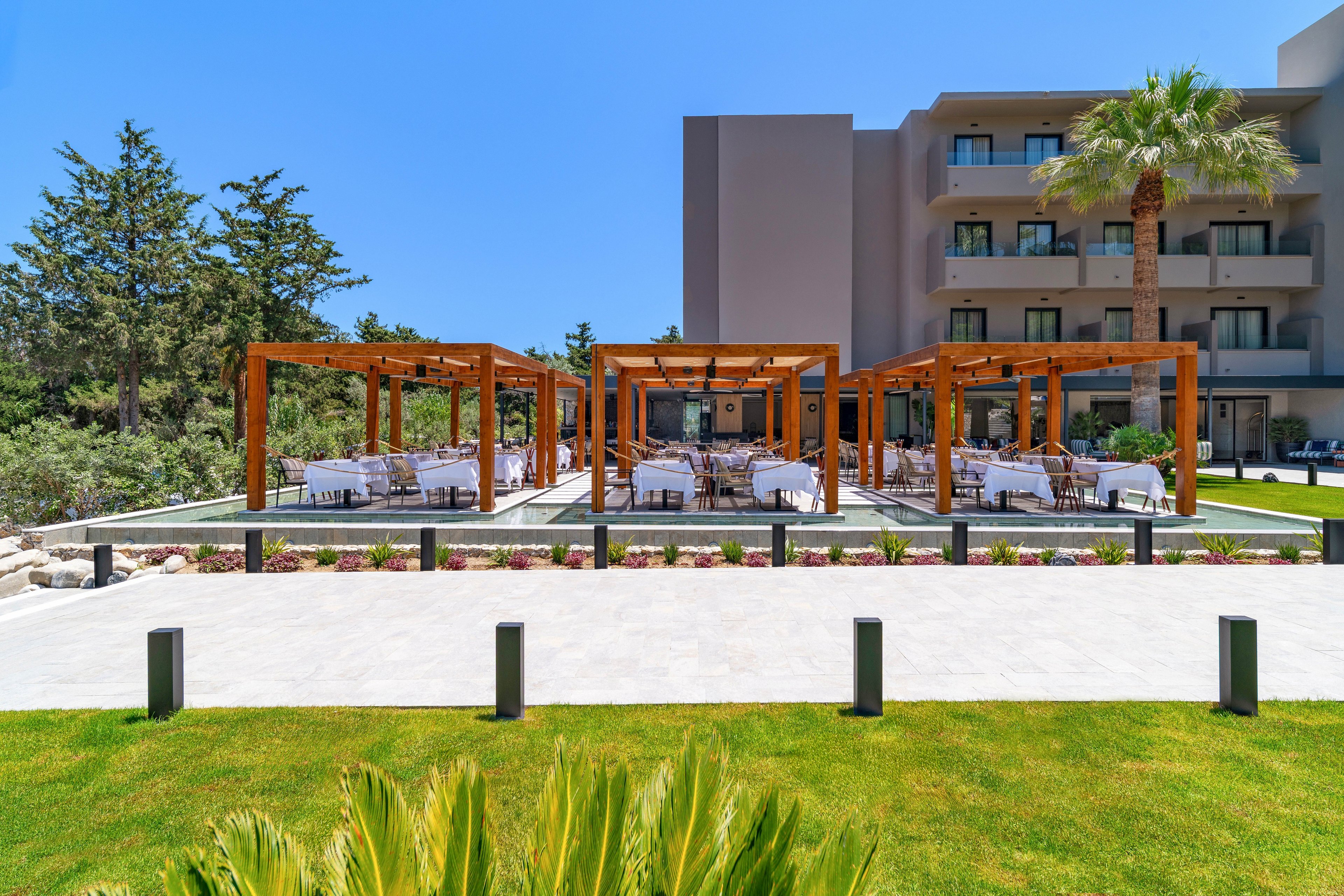 An outdoor dining terrace is set up in front of a modern hotel building. Multiple wooden pergolas shelter neatly arranged tables and chairs, creating an inviting alfresco seating area. The foreground features a manicured lawn, paved walkway, and low bollard lights. Trees and greenery surround the space under a clear blue sky, suggesting a warm, sunny day.