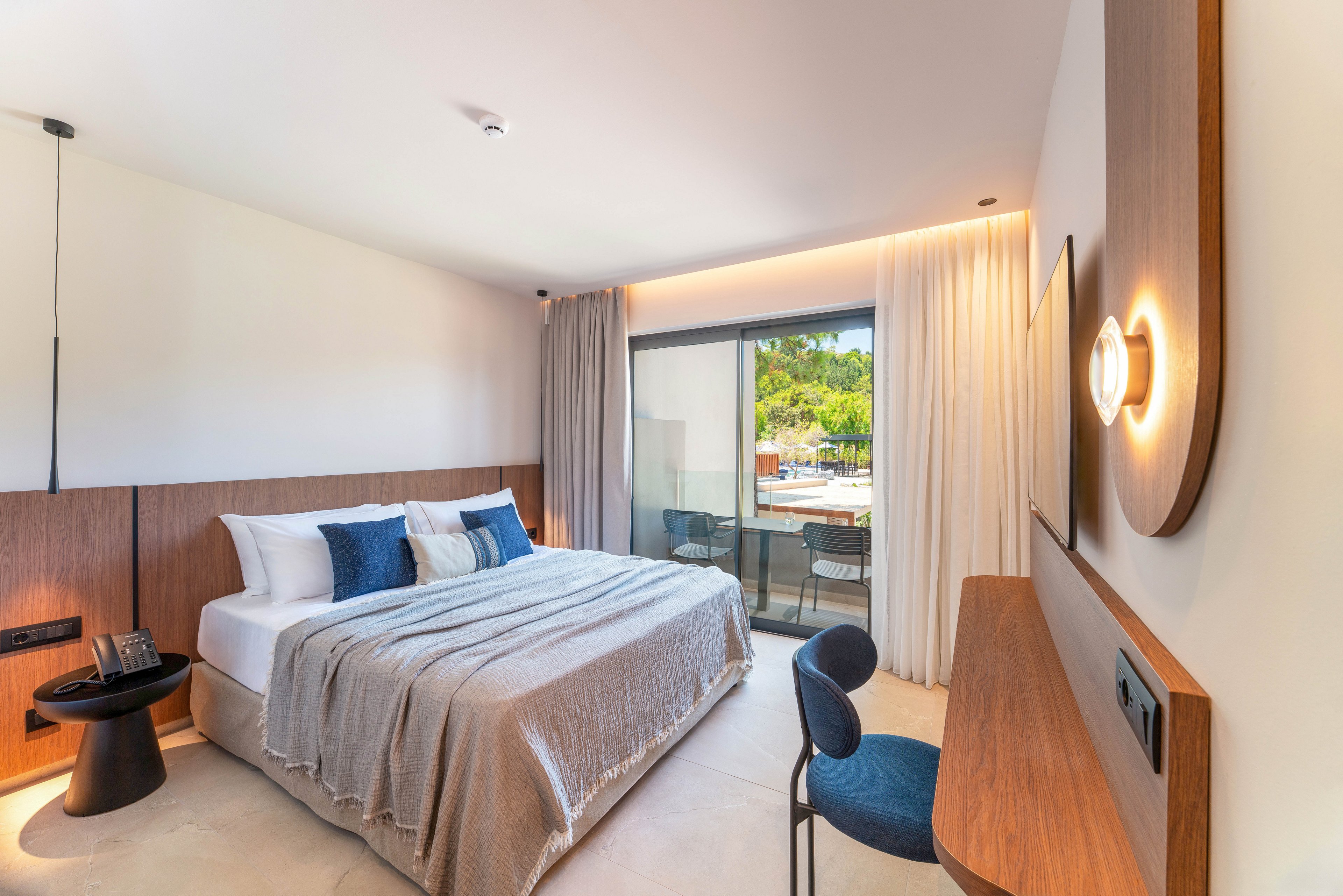A contemporary hotel bedroom features a neatly made double bed with neutral bedding and blue accent pillows. Warm wooden paneling and furniture create a minimalist, cozy atmosphere. A sliding glass door opens to a balcony with outdoor seating and a view of greenery and a pool area. Soft ambient lighting and sheer curtains enhance the bright, inviting space.