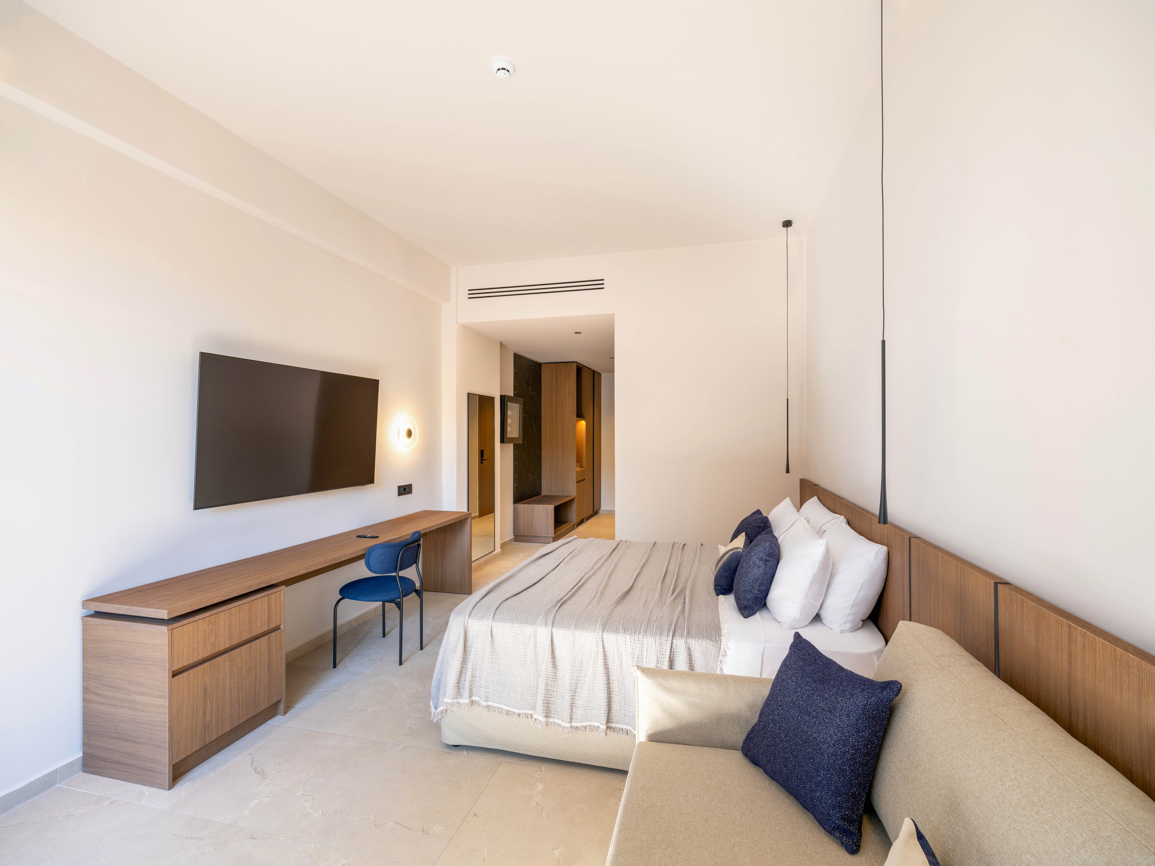 A bright modern hotel room features a neatly made bed with multiple pillows and a light bedspread. A large flat-screen television is mounted above a long wooden desk with a blue chair. In the foreground, a beige sofa with dark blue cushions adds extra seating. Neutral walls and flooring, along with simple lines and minimal decor, create a clean and contemporary atmosphere.