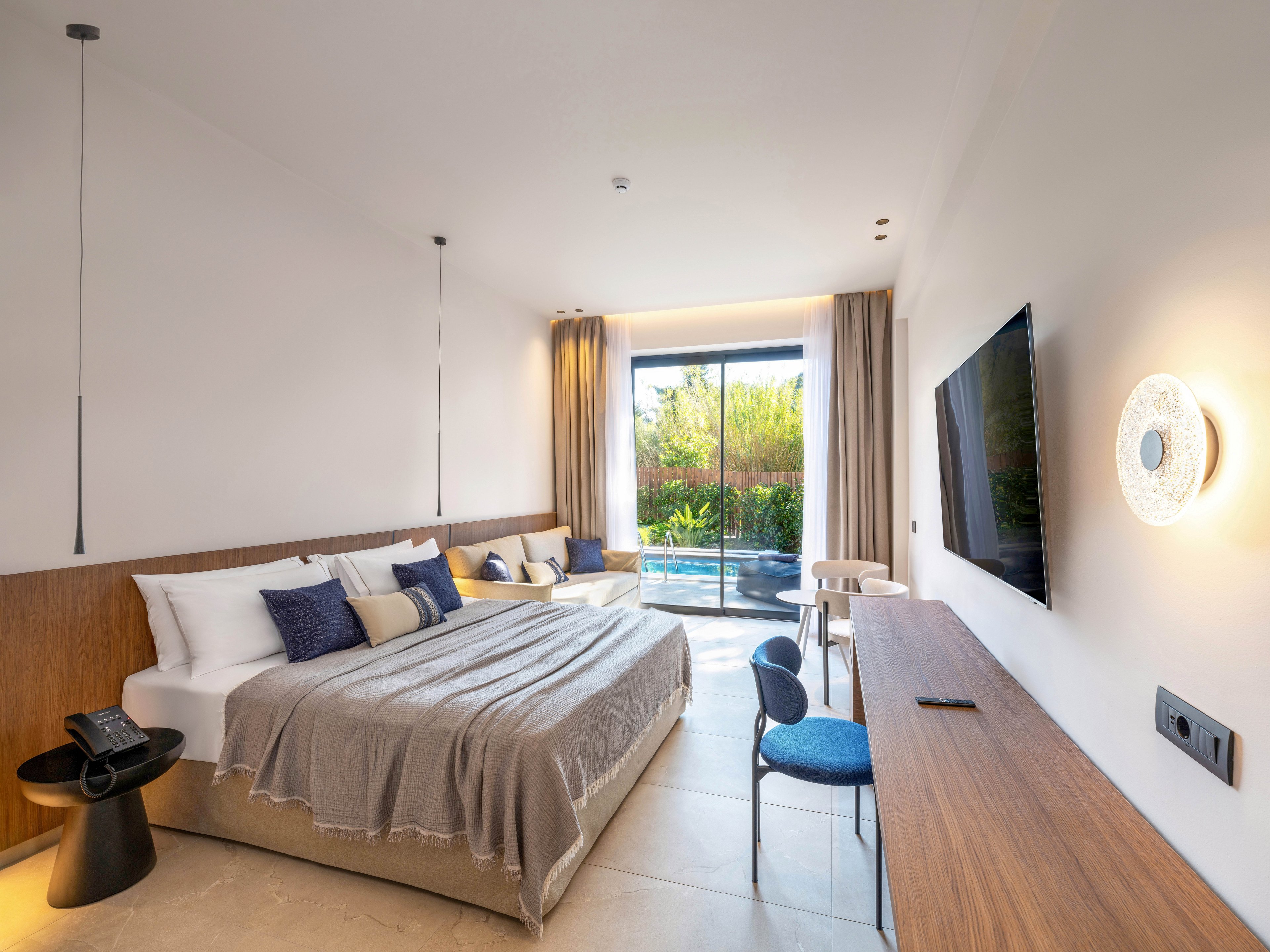 A contemporary hotel bedroom features a large bed with neutral bedding and multiple accent pillows. A long wooden desk runs along the wall opposite a mounted flat-screen television. Floor-to-ceiling glass doors open to a patio with a small private pool and lush greenery outside. Minimalist lighting, light-colored walls, and simple furnishings create a calm, airy atmosphere.