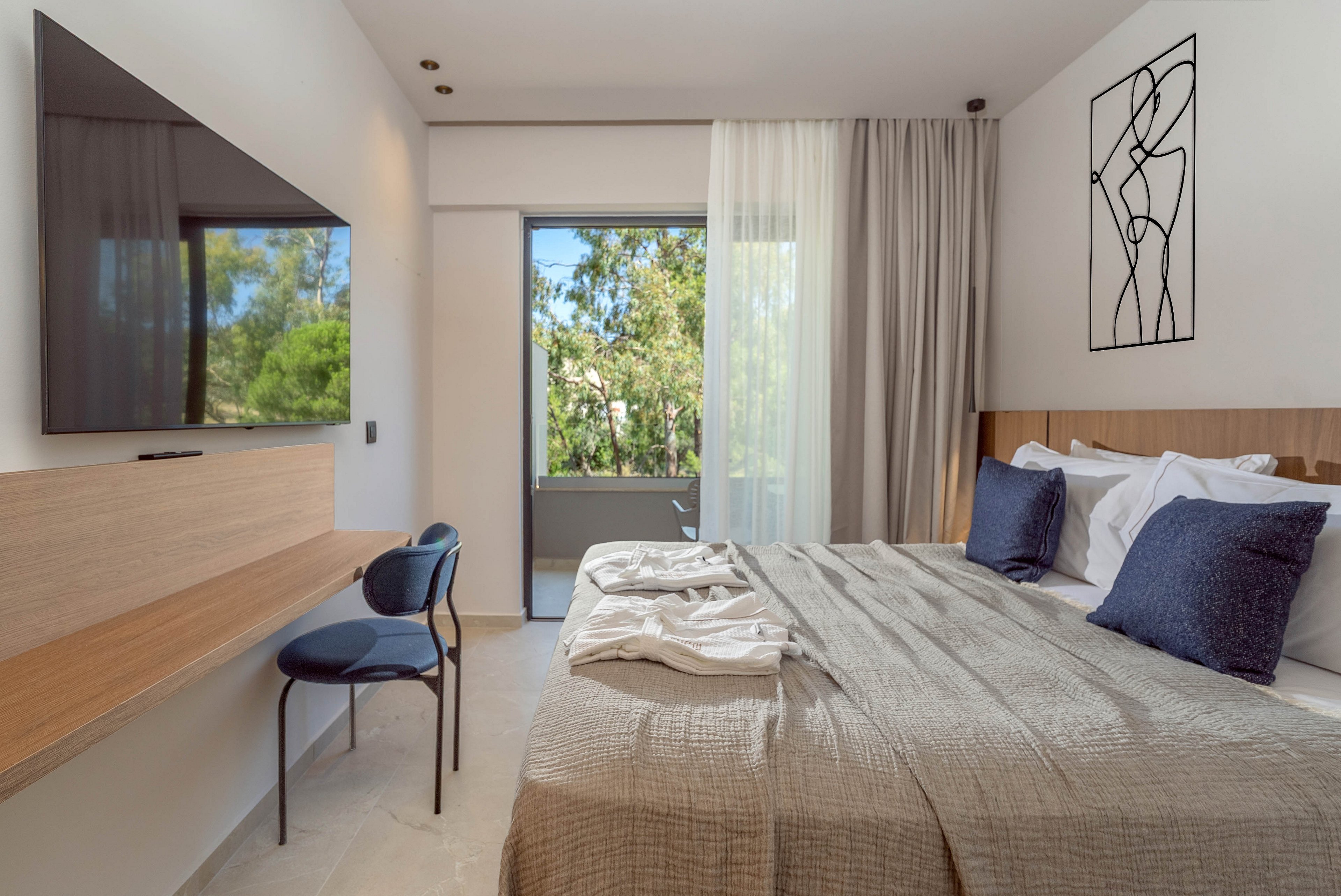 A contemporary bedroom features a neatly made bed with neutral-toned bedding and accent pillows. A large flat-screen television is mounted on the wall above a slim wooden desk with a dark chair. Sheer curtains frame a glass door that opens to a balcony with lush green trees outside. The room is decorated in a minimalist style with clean lines, light walls, and a simple abstract wall art piece.