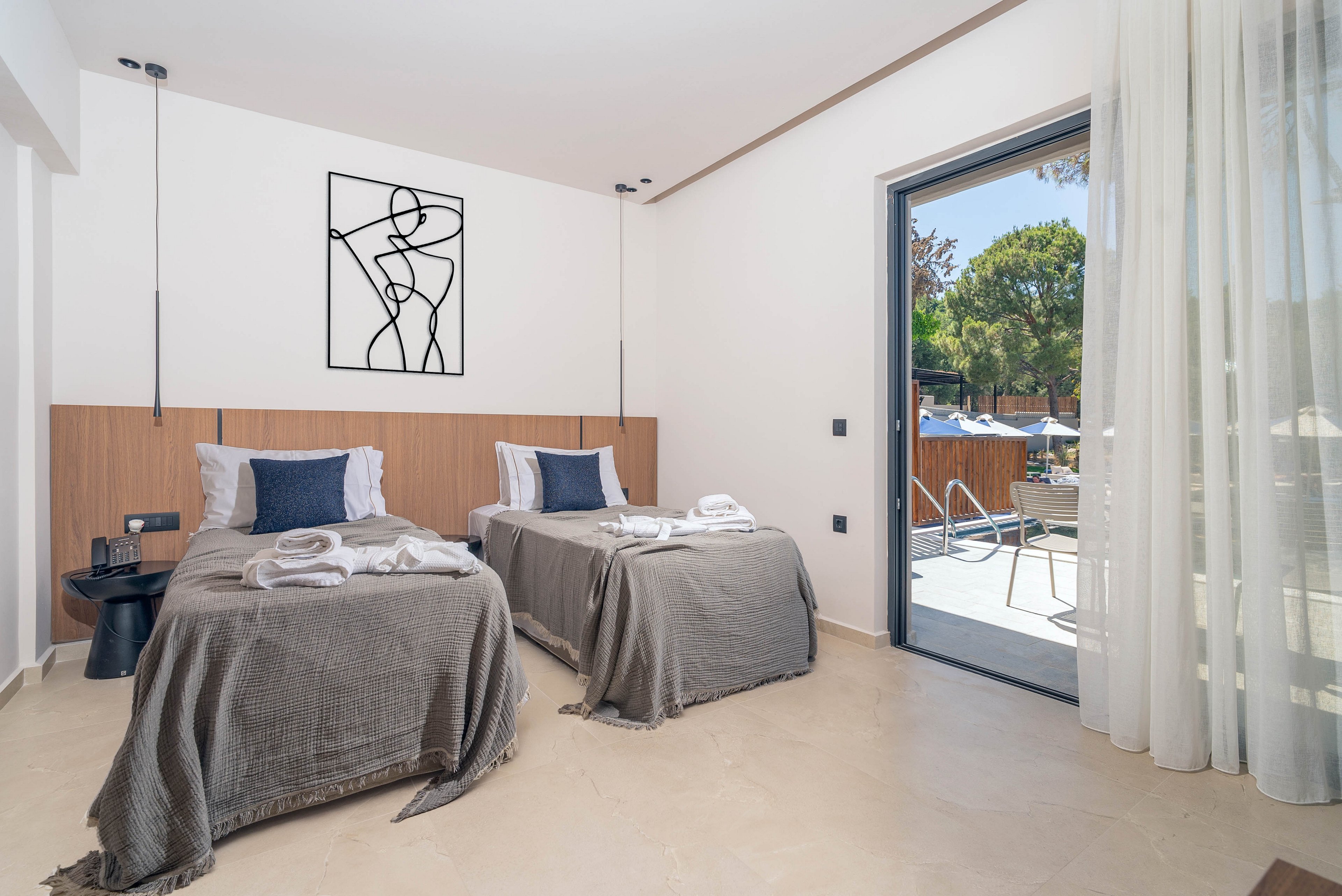 A contemporary hotel room features two neatly made twin beds with gray throws and dark blue accent pillows. The space is bright and minimal, with light-colored floors, a wooden headboard panel, and a black line-art wall piece above the beds. A sliding glass door opens to a sunny balcony with outdoor seating and views of trees and a pool area. Sheer curtains and subtle lighting create an airy, relaxed atmosphere suitable for vacation or business travel.