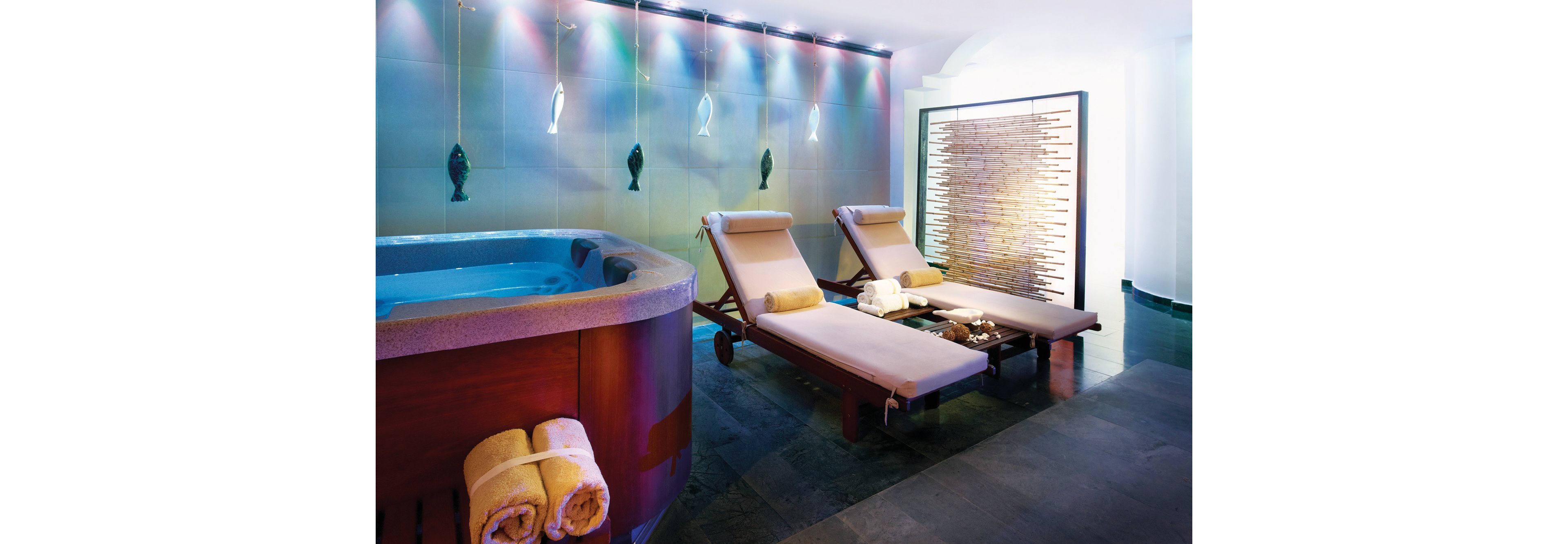 A serene spa setting featuring a wooden hot tub with rolled towels and two reclining lounge chairs. The room is illuminated with soft, colorful lighting and decorative fish wall accents. A textured partition adds privacy and style to the tranquil atmosphere.