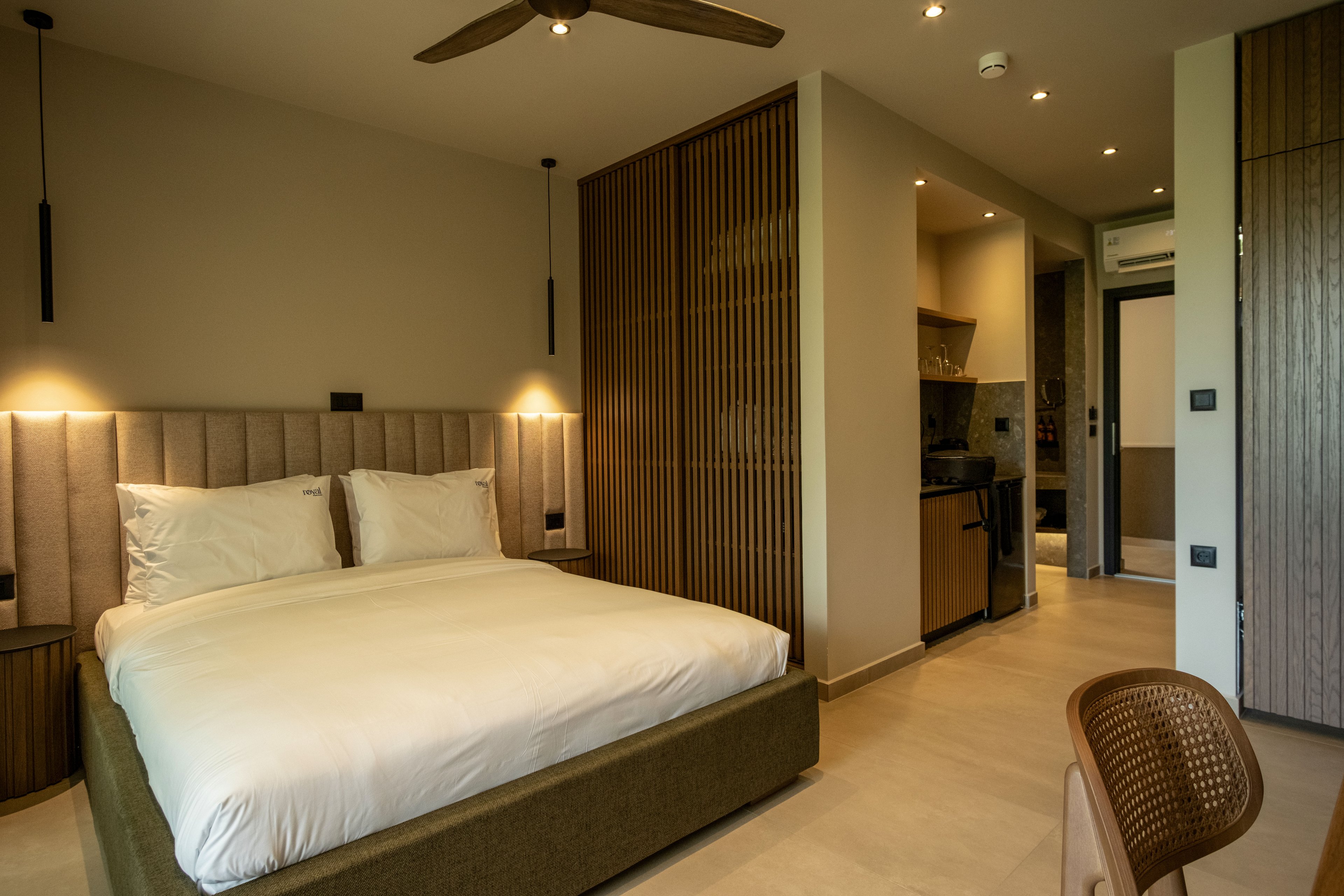 Contemporary studio-style hotel room featuring a large bed with white linens and upholstered headboard. The space includes warm wood accents, vertical slatted partition, and recessed ceiling lights. A compact kitchenette with dark countertop and cabinets is visible in the background. Neutral tones and minimal decor create a calm, cozy atmosphere suitable for short stays or vacation rentals.