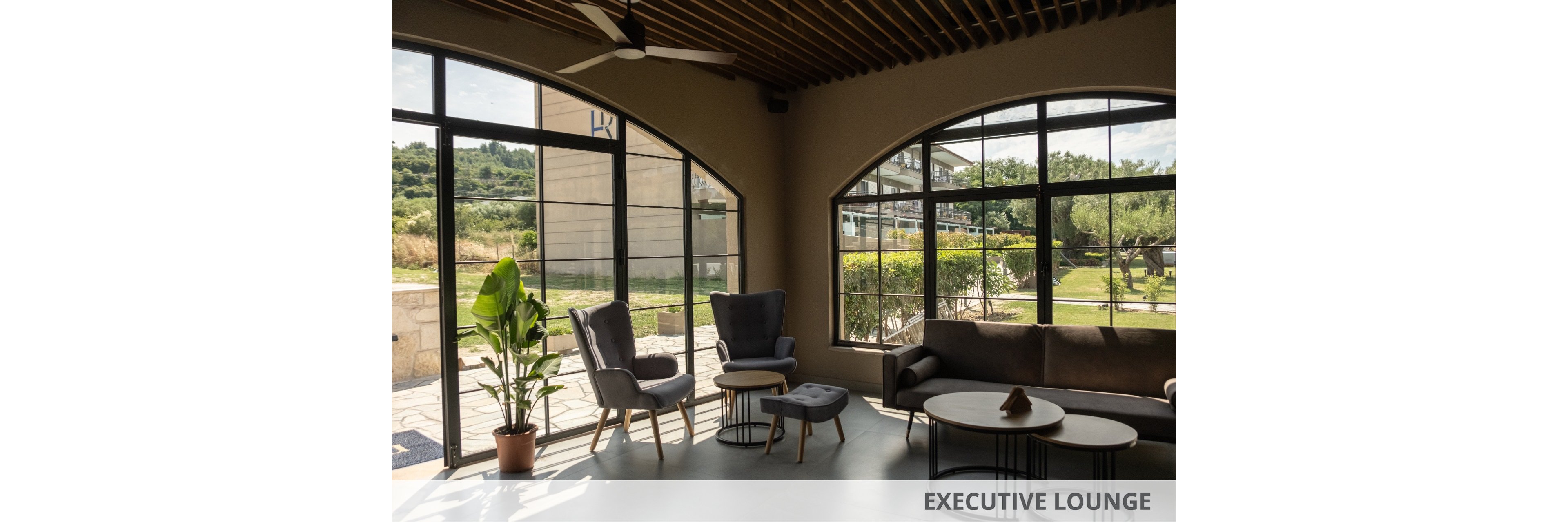 A contemporary lounge area features a mix of armchairs, a sofa, and nested coffee tables arranged near expansive arched windows. Natural light floods the room, highlighting the neutral color palette and minimalist decor. A potted plant adds greenery indoors while the view outside shows a landscaped garden and surrounding buildings. The space feels calm and inviting, suitable for relaxation or informal meetings.