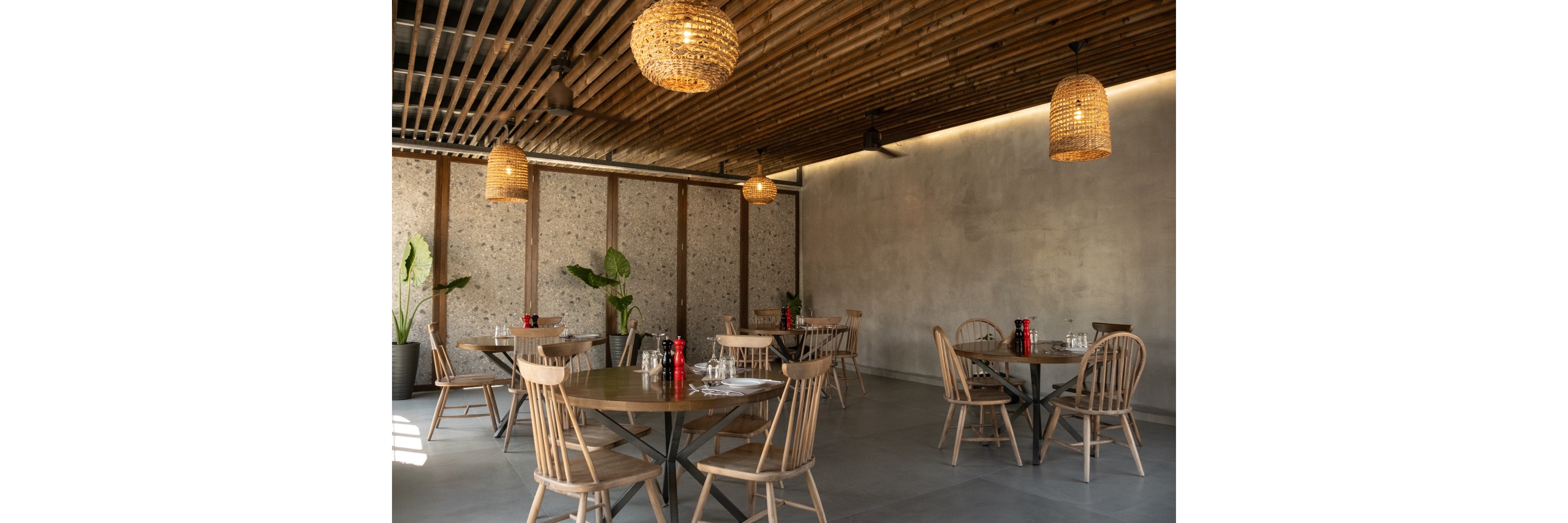 Contemporary restaurant dining area featuring round wooden tables and simple wooden chairs. The space has a minimalist design with concrete walls, large floor tiles, and warm wooden ceiling slats. Woven pendant lights create a cozy ambiance above the tables, which are set with glassware and condiments. Potted plants along the wall add a touch of greenery to the calm, modern setting.