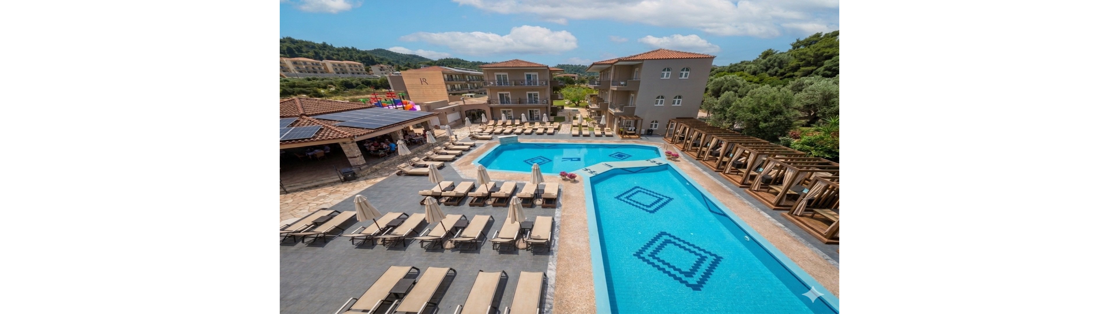 A large outdoor swimming pool complex is surrounded by numerous beige lounge chairs and umbrellas at a resort. Several low-rise hotel buildings frame the pool area, set against a backdrop of green hills and trees. The bright blue water features geometric tile patterns, and a clear sky with scattered clouds enhances the summery atmosphere. Wooden cabanas line one side of the pool, creating a relaxing vacation setting.
