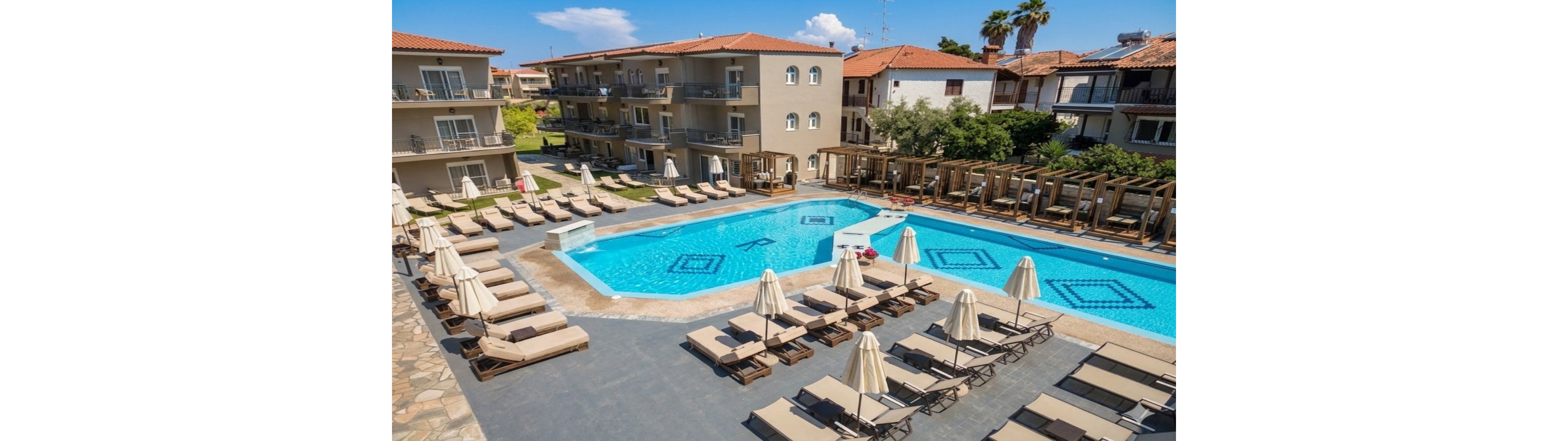 A modern resort courtyard features two bright blue outdoor swimming pools surrounded by sun loungers and umbrellas. Low-rise hotel buildings with balconies enclose the pool area, creating an intimate vacation setting. Wooden cabanas line one side of the pool deck, offering shaded relaxation spaces. The scene is sunlit and calm, suggesting a warm, inviting holiday atmosphere.