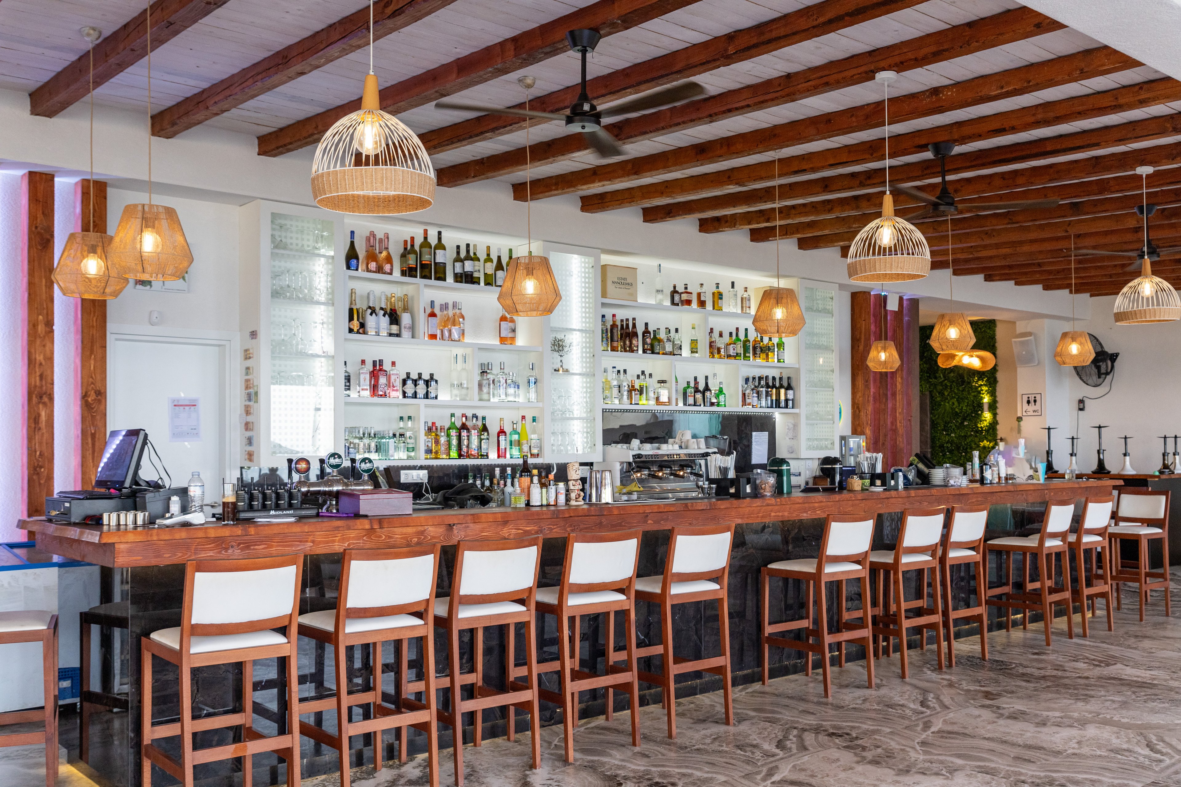 Contemporary bar interior featuring a long wooden counter lined with high stools. The back wall is filled with shelves of assorted liquor bottles and glassware. Warm wicker pendant lights hang from a ceiling with exposed wooden beams, creating a relaxed tropical atmosphere. The space is bright and airy, with neutral flooring and clean white walls emphasizing the casual resort-style setting.