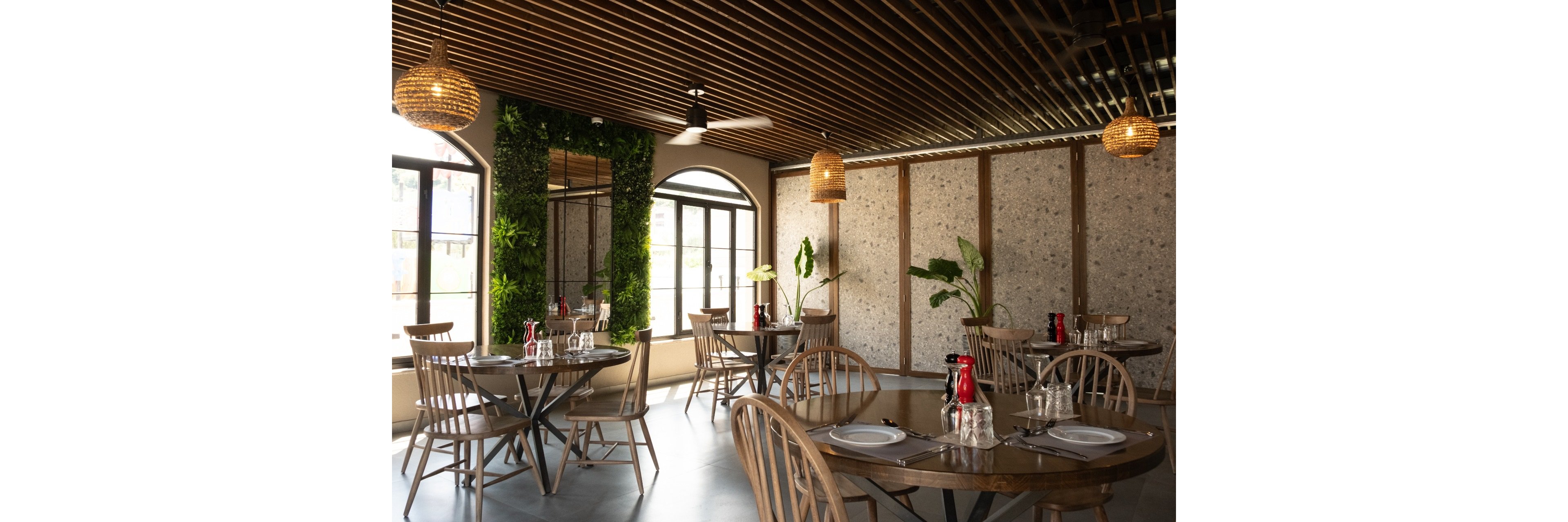 Contemporary restaurant dining area featuring round wooden tables and spindle-back chairs. The space is bright with large arched windows and a wall of vertical greenery adding a natural touch. Warm pendant lights and a slatted wooden ceiling create a cozy, inviting atmosphere. Tables are neatly set with plates, glasses, cutlery, and red water bottles, ready for guests.