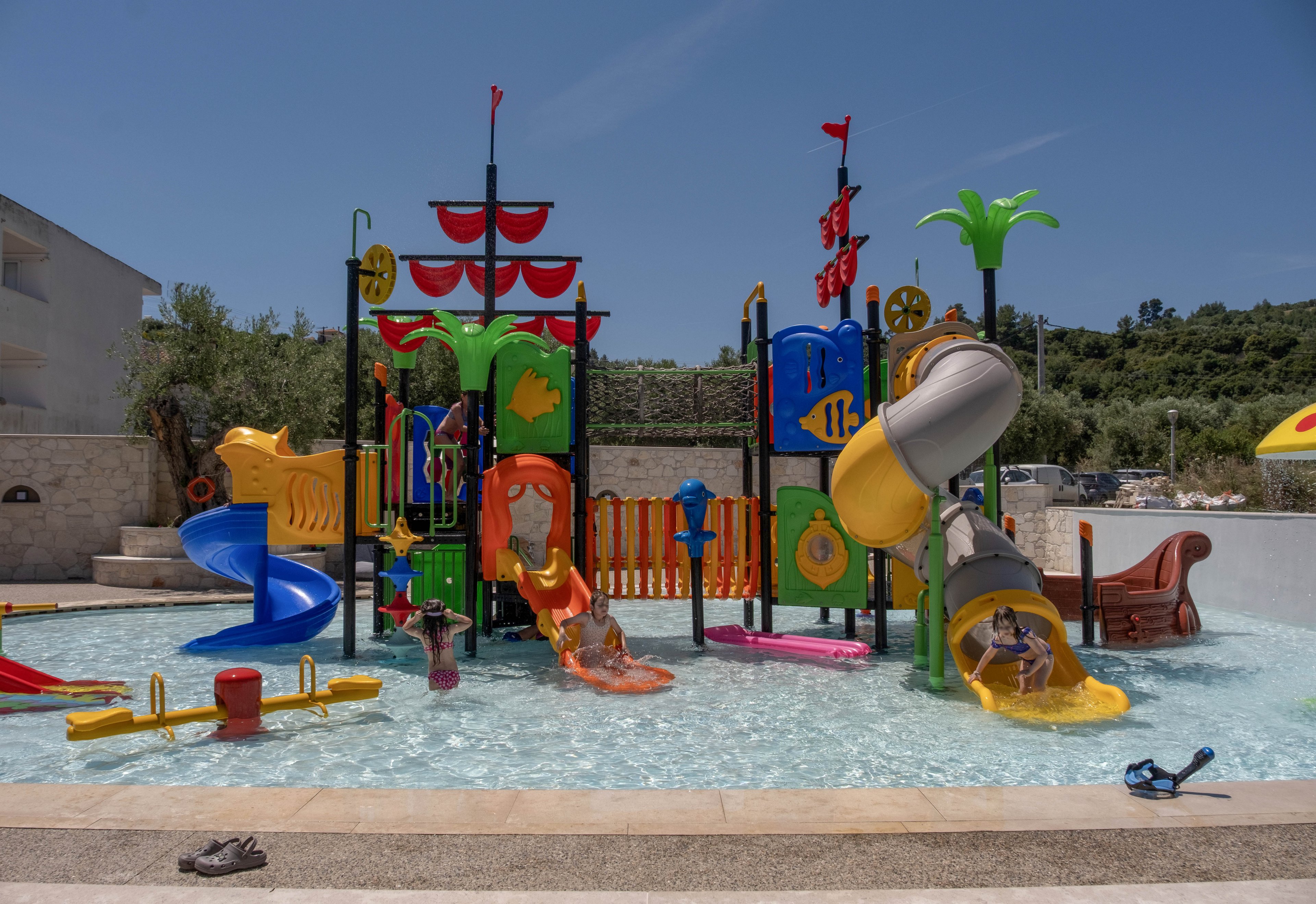 Children play on a colorful pirate-themed water playground in a shallow outdoor pool. The structure features multiple slides, climbing elements, and water sprayers in bright red, yellow, blue, and green. Surrounding the play area are low walls, greenery, and clear blue sky, suggesting a warm summer day. The scene feels fun, energetic, and family-friendly.