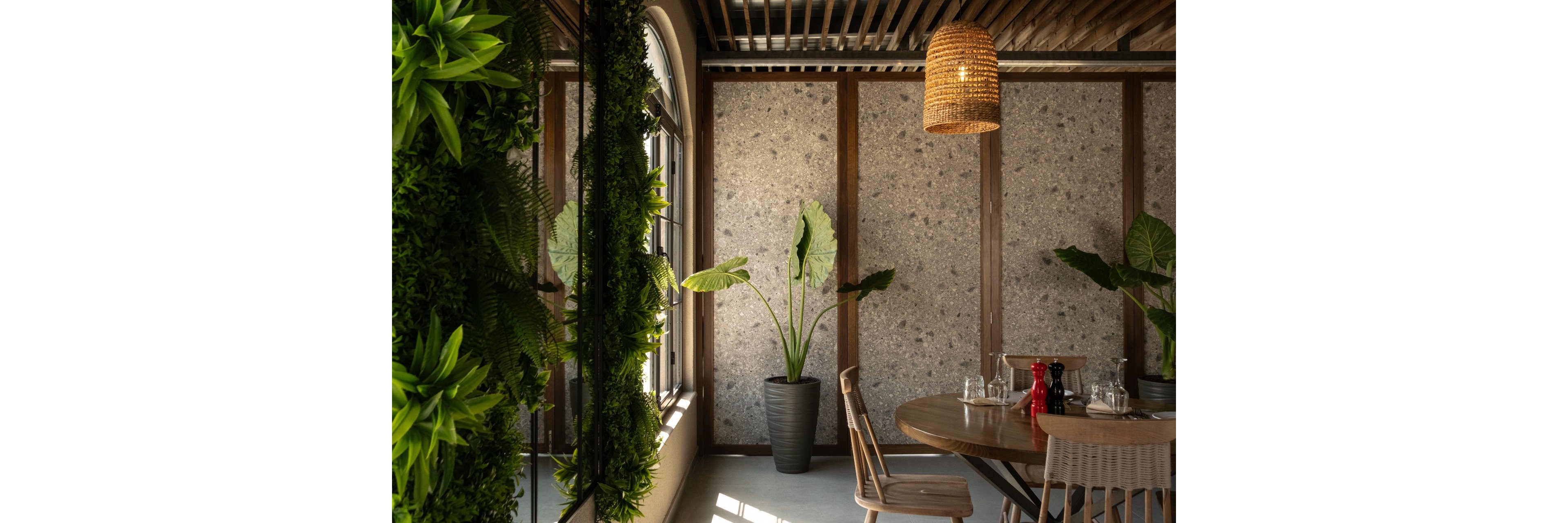A small dining table is set in a modern interior corner with natural light streaming through a nearby window. The space features a round wooden table with glassware and a red pepper mill, surrounded by simple wooden chairs. Large potted plants and a vertical wall of greenery add a lush, biophilic touch. A woven pendant light and exposed ceiling elements contribute to the warm, contemporary atmosphere.