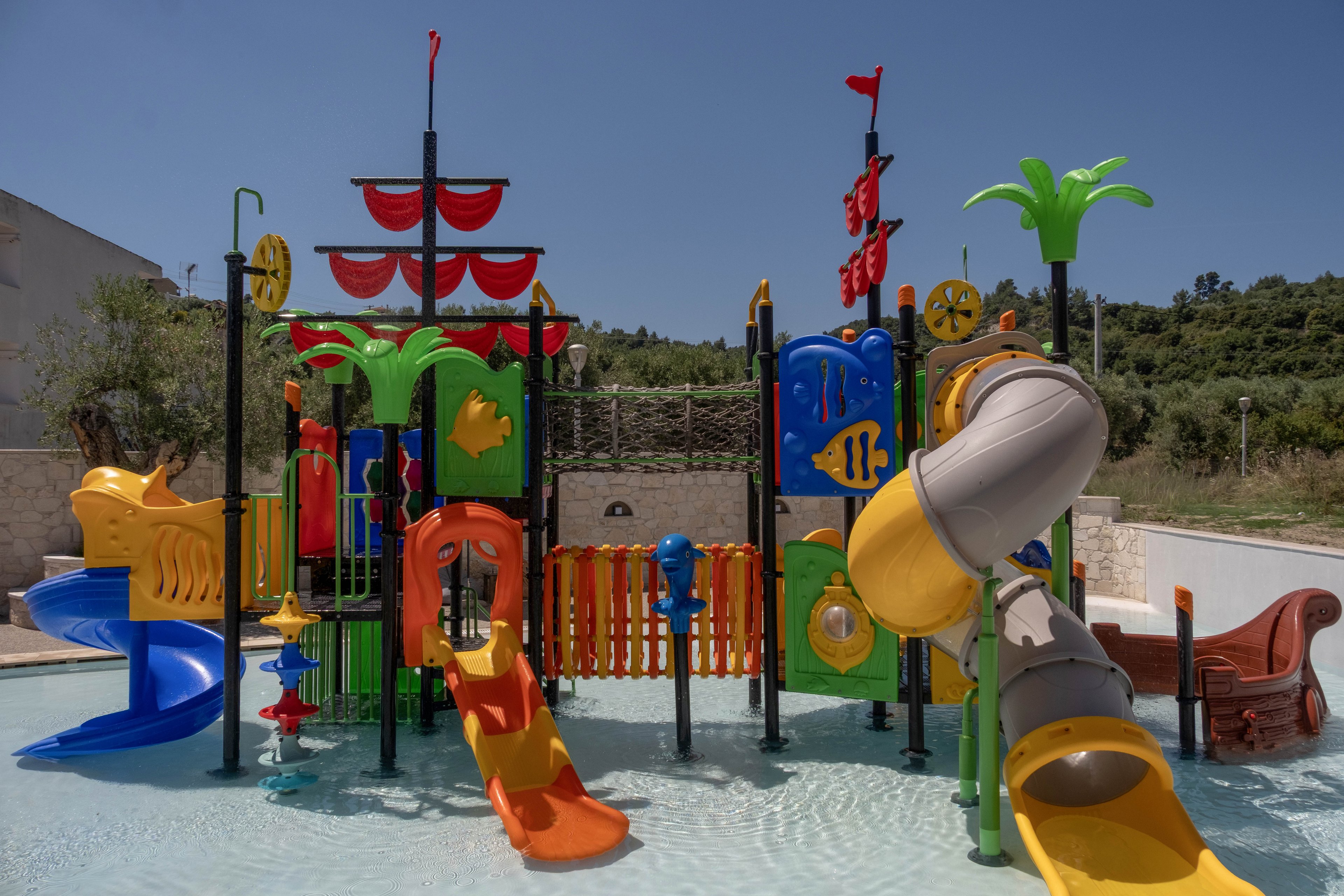 A brightly colored water playground structure stands in a shallow pool under a clear blue sky. Multiple slides, climbing elements, and tunnels create a fun aquatic play area for children. The design features nautical and animal shapes, including fish, palm trees, and a ship-like section. The surrounding area includes stone walls and greenery, suggesting an outdoor resort or water park setting.