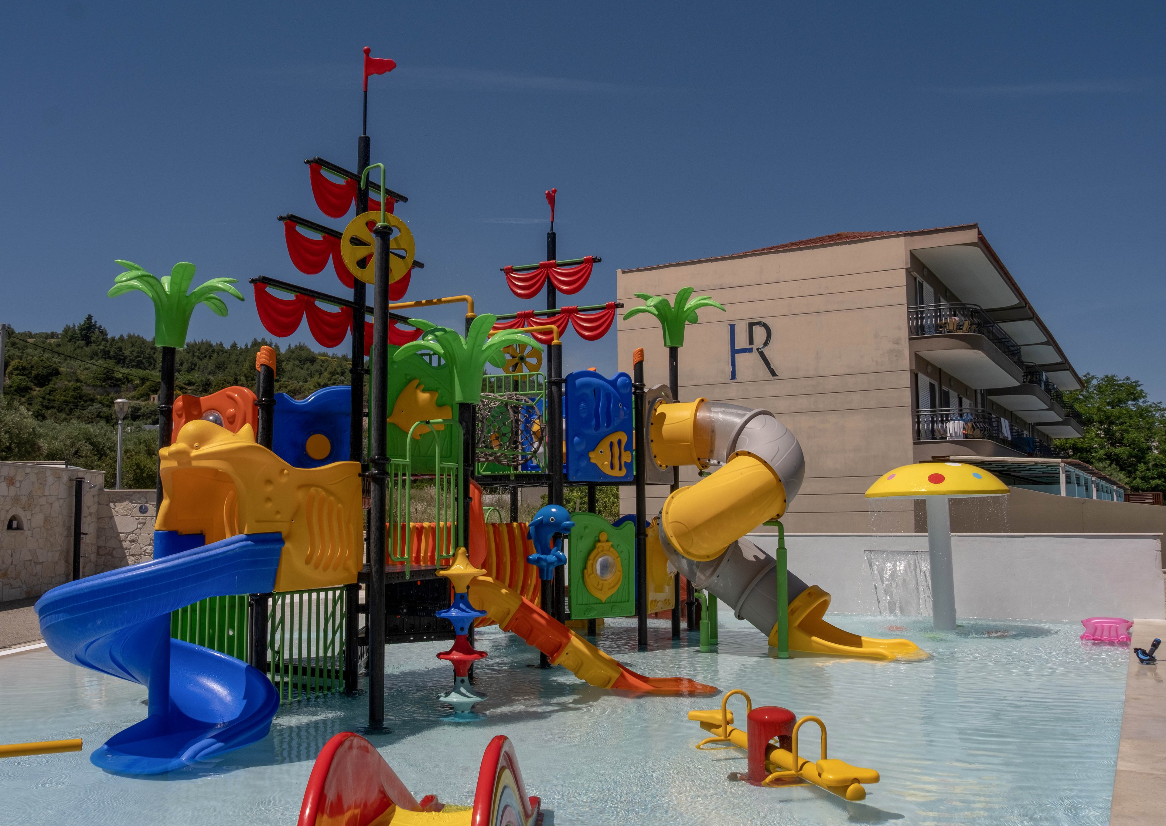 A vibrant water playground with slides and climbing structures stands in a shallow outdoor pool. The play area features bright yellow, red, blue, and green elements, including palm tree decorations and a spiral tube slide. In the background, a modern resort building with balconies and the letters "LR" on the wall is visible. The scene is lit by clear daylight under a blue sky, creating a fun and inviting atmosphere for children.