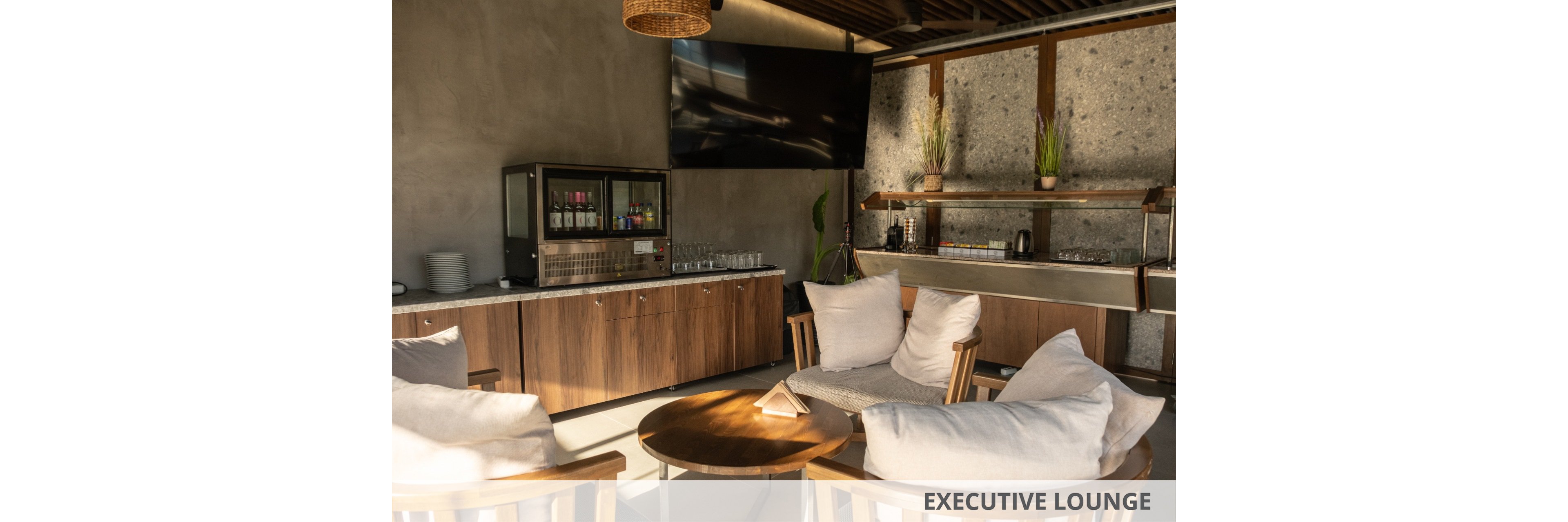 A contemporary lounge area features wooden chairs with white cushions arranged around a small round table. In the background, a beverage station with a glass-front cooler and neatly stacked glasses sits against a concrete wall. A large flat-screen television is mounted above the counter, and a secondary counter with plants and shelves adds to the decor. Warm natural light and neutral tones create a relaxed, modern atmosphere.