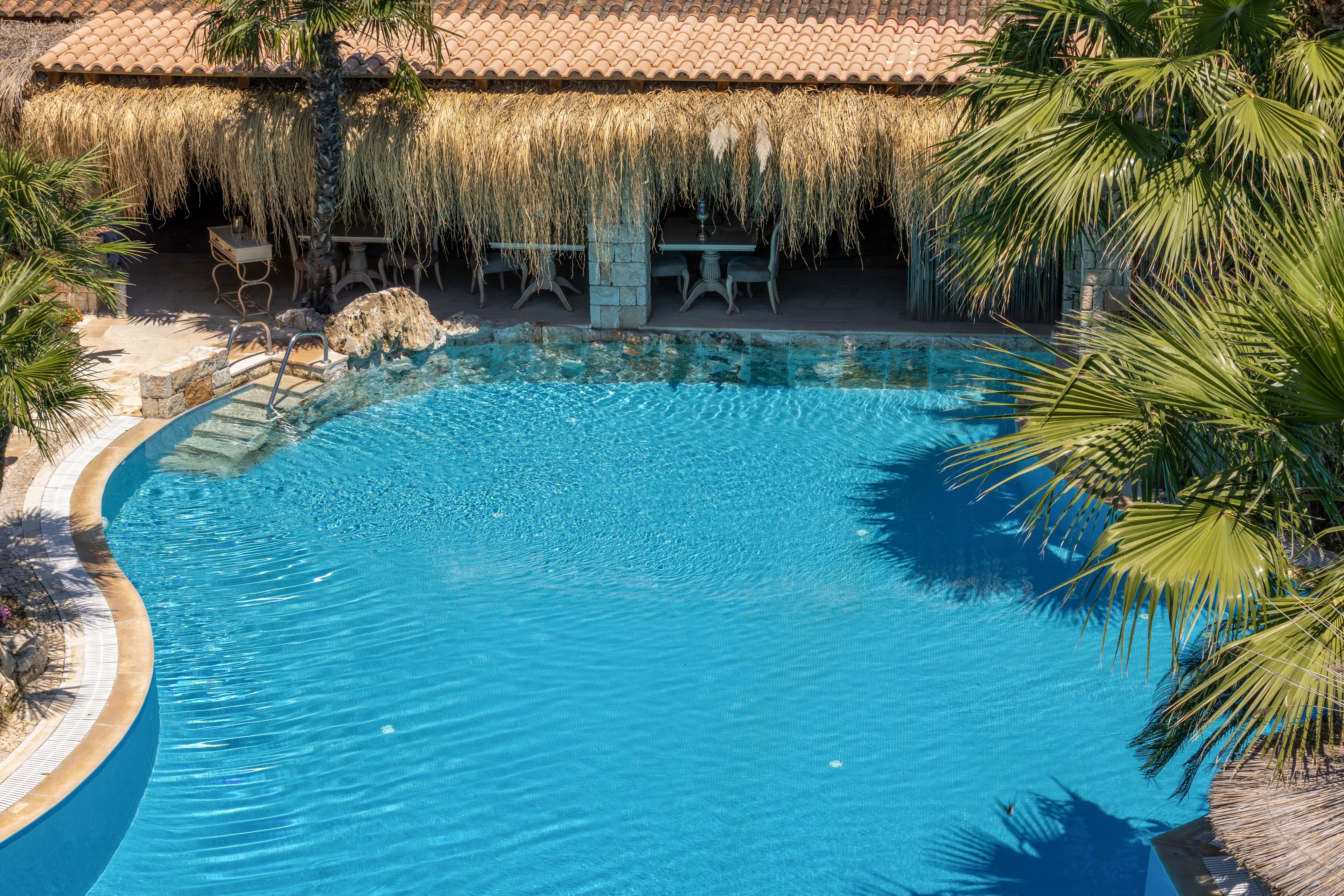 A clear blue swimming pool curves around a tropical-style bar area with a thatched roof. Palm trees frame the water, creating a lush, vacation atmosphere. Bar stools and tables sit under the shaded structure, suggesting a relaxed poolside dining or drink setting. The bright sunlight and reflections on the water emphasize a warm, inviting holiday mood.