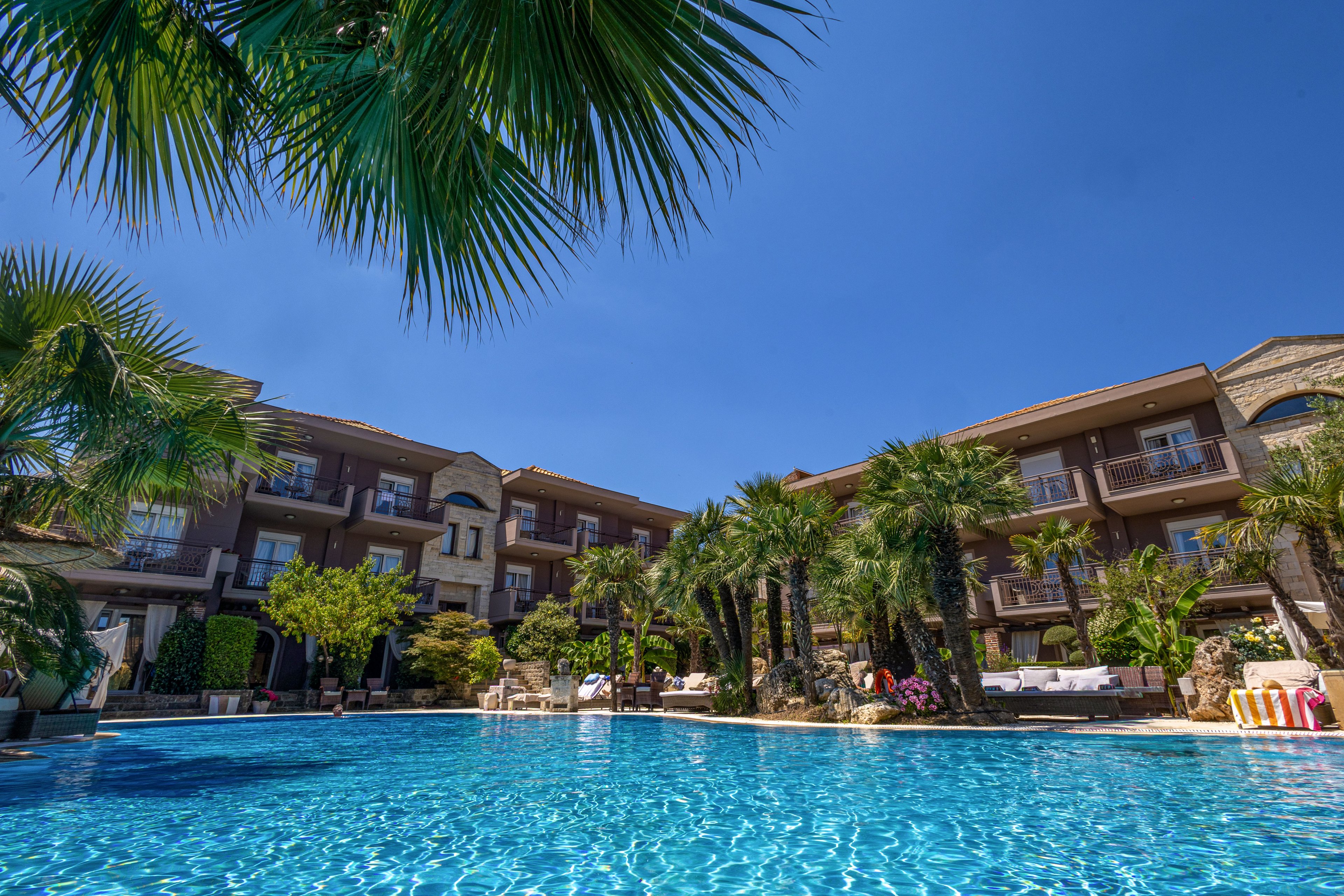 A large outdoor swimming pool stretches across the foreground of a luxury resort hotel. The multi-story building features balconies, stone accents, and lush greenery surrounding the pool area. Tall palm trees and tropical plants create a vacation atmosphere under a clear blue sky. Sun loungers and seating areas along the pool edge suggest a relaxing holiday setting.