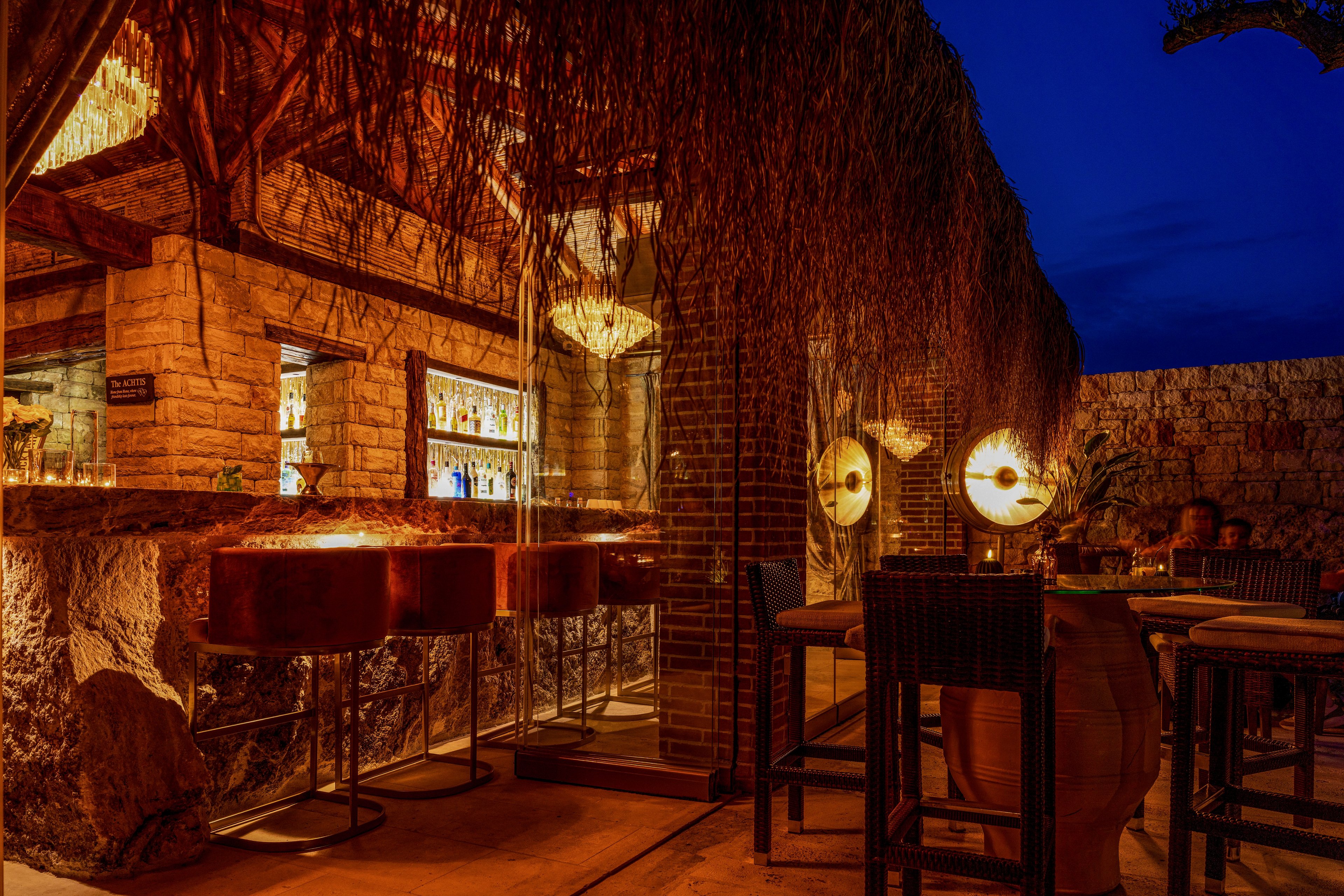 An inviting outdoor bar is illuminated with warm golden lights against rustic stone and wood surfaces. High stools line the bar counter, while nearby tables and chairs create an intimate seating area. Decorative hanging lamps and textured thatched elements add to the cozy atmosphere. The deep blue night sky contrasts with the amber glow, emphasizing a relaxed, upscale nightlife setting.