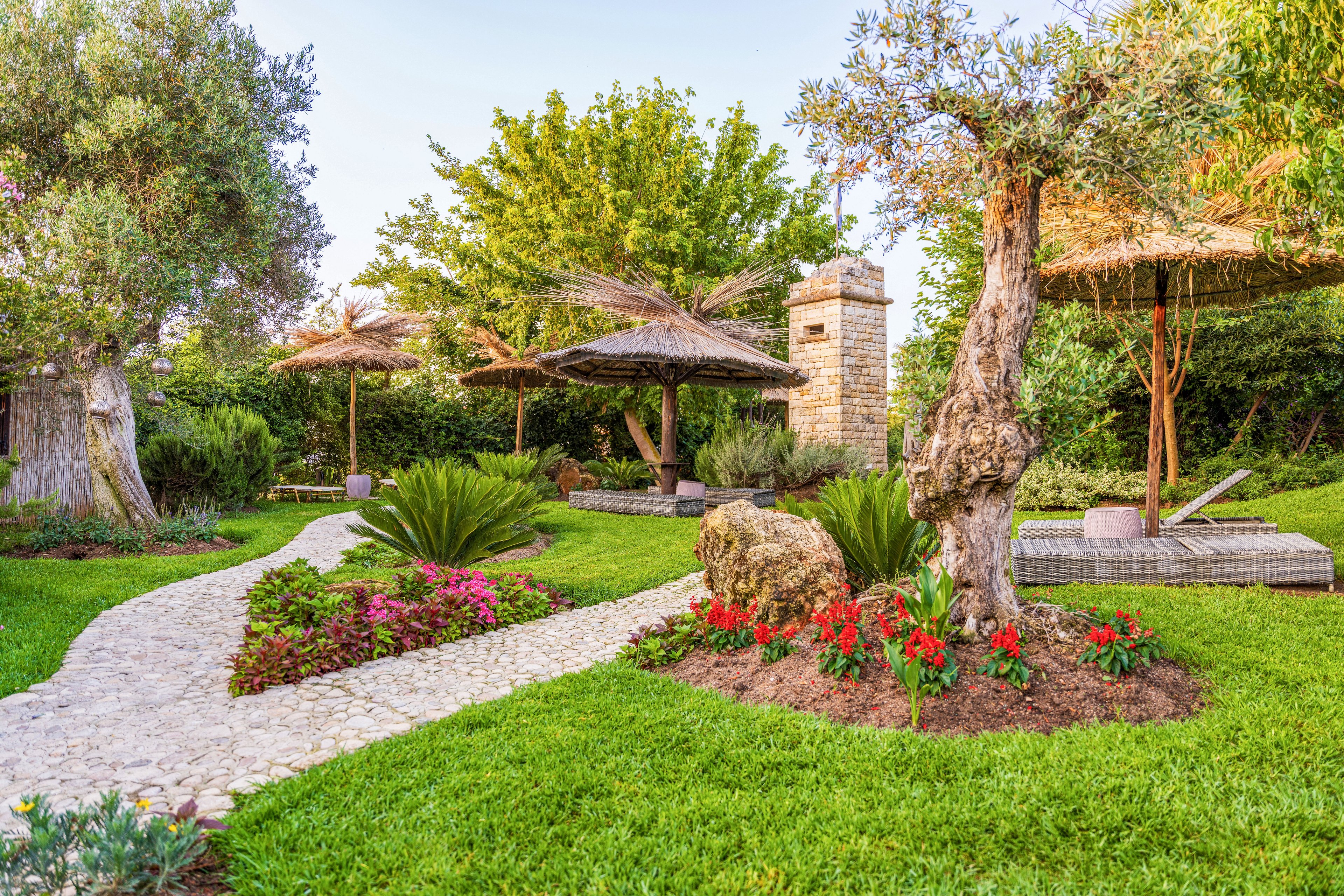 A lush landscaped garden features a curving stone pathway leading through vibrant green grass and colorful flower beds. Palm-style thatched umbrellas and wicker lounge furniture create shaded seating areas in the background. Mature trees, ornamental shrubs, and a large rock add texture and structure to the scene. The overall setting feels peaceful and inviting, ideal for outdoor relaxation in warm weather.