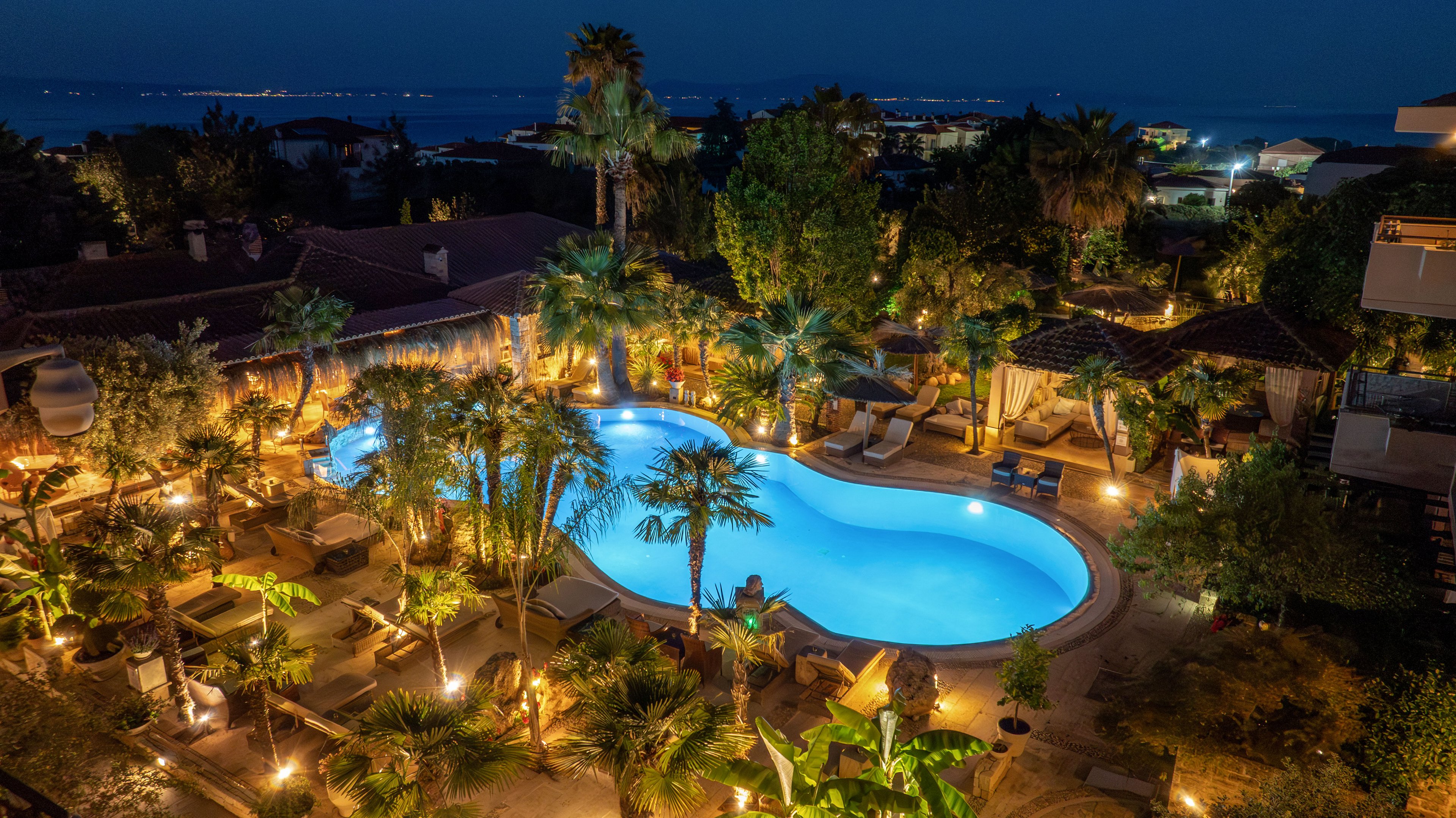 An outdoor hotel swimming pool is brightly lit at night, surrounded by lush palm trees and garden landscaping. Lounge chairs and cabanas are arranged around the curved pool, creating a relaxing resort atmosphere. Warm ambient lighting highlights the pathways and seating areas, while nearby buildings frame the courtyard. In the distance, faint lights along the coastline suggest a seaside location under a dark blue sky.